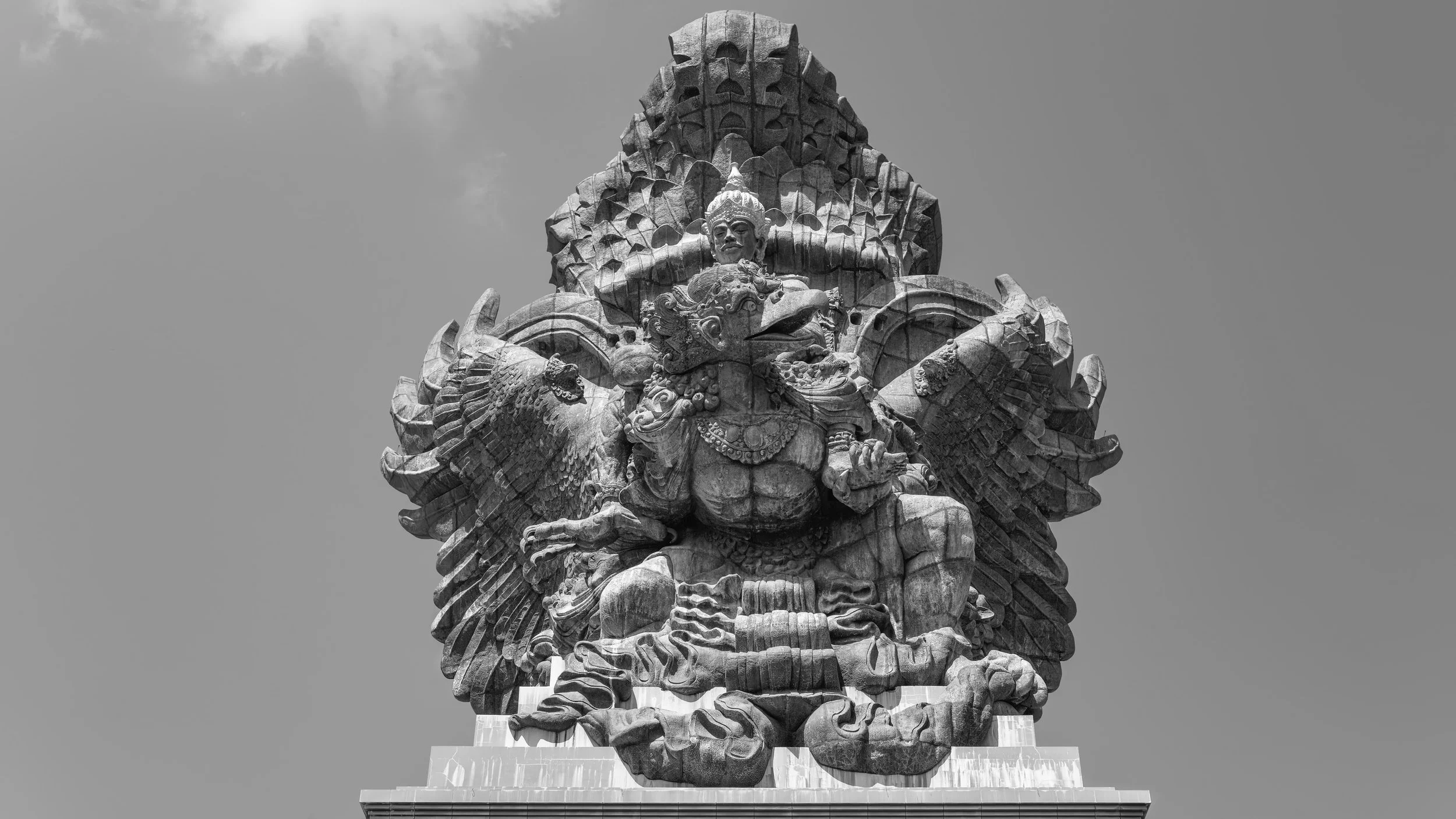 Black and white photograph of a large stone statue depicting a fierce, multi-armed deity with a crown and ornate clothing, seated on a base against a cloudy sky.