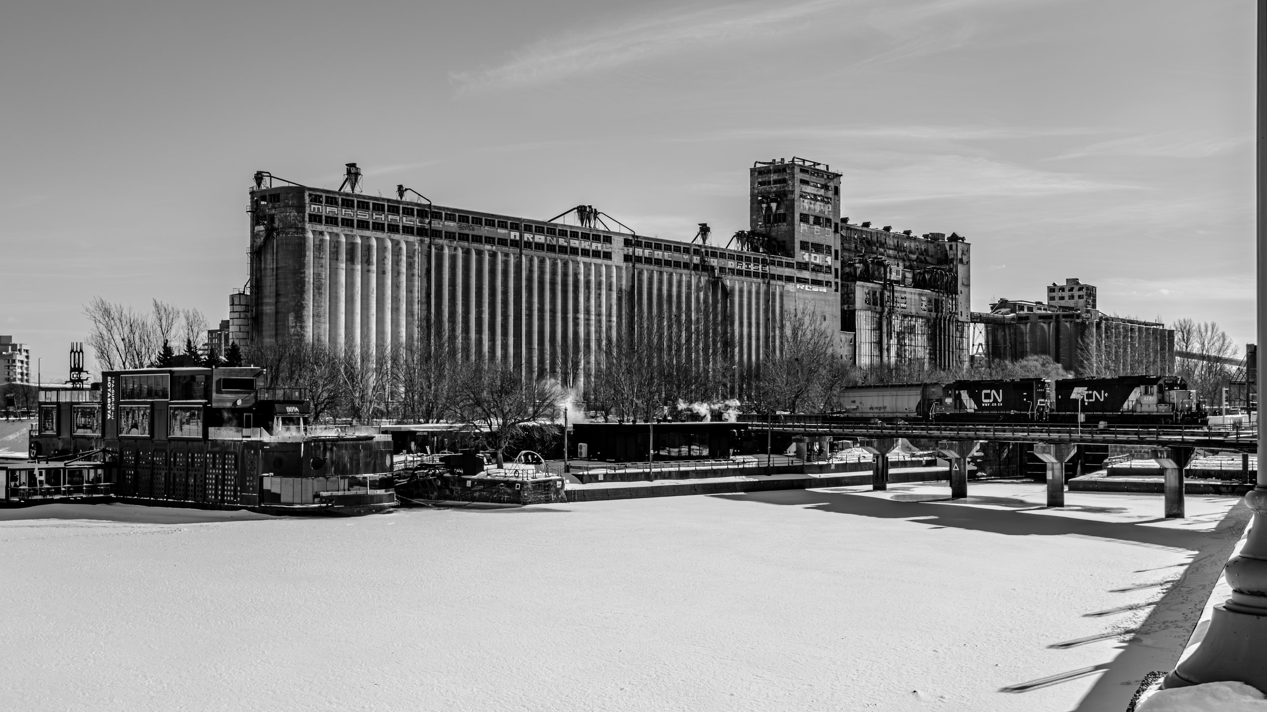 Black and white photo of an industrial building, likely a grain elevator or silo, with train tracks and locomotives in front, winter scene with snow-covered ground and trees.