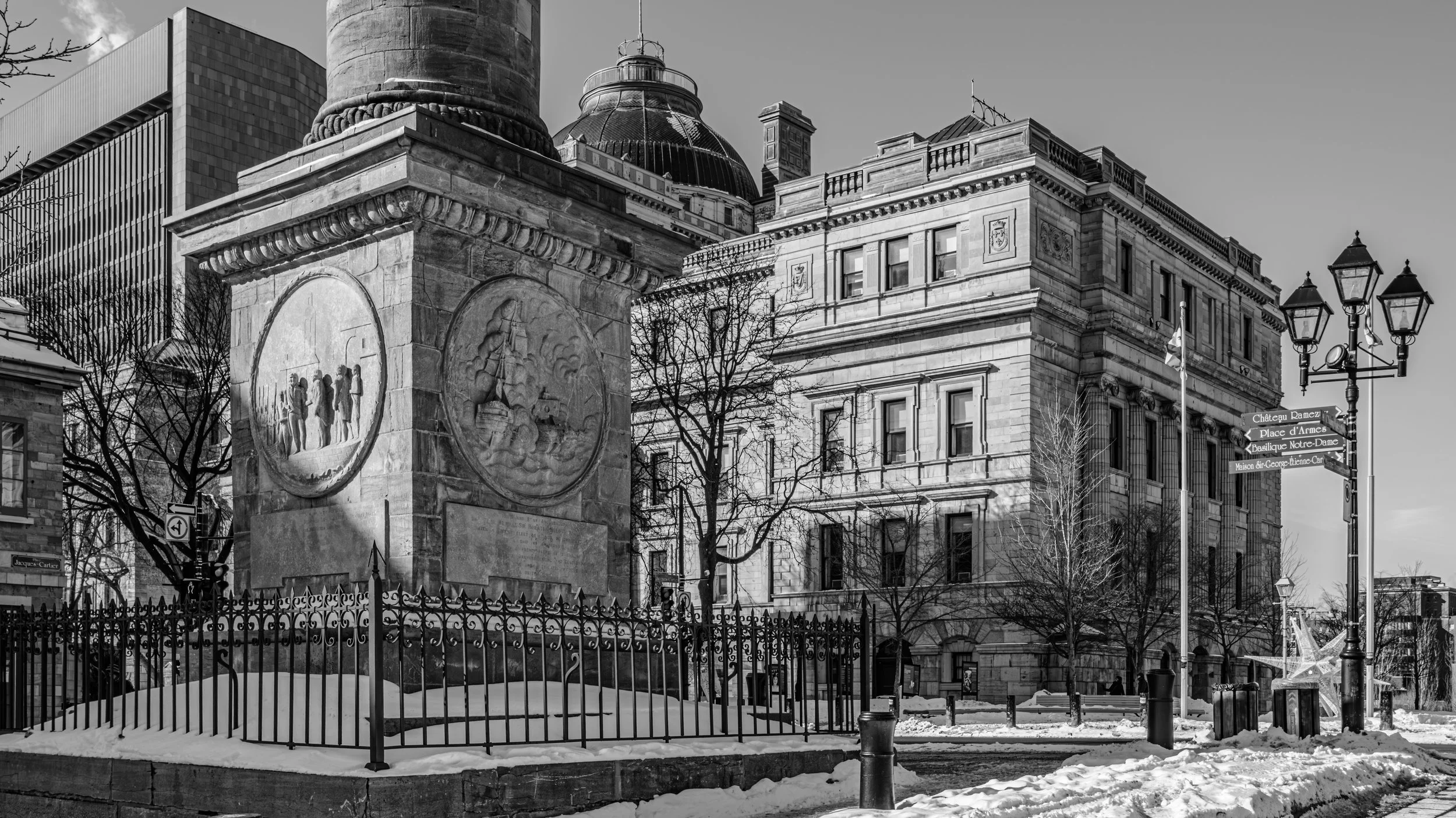 Black and white photo of a historic stone monument with carved reliefs, surrounded by a decorative metal fence, in a city scene with classical and modern buildings, leafless trees, and street signs.