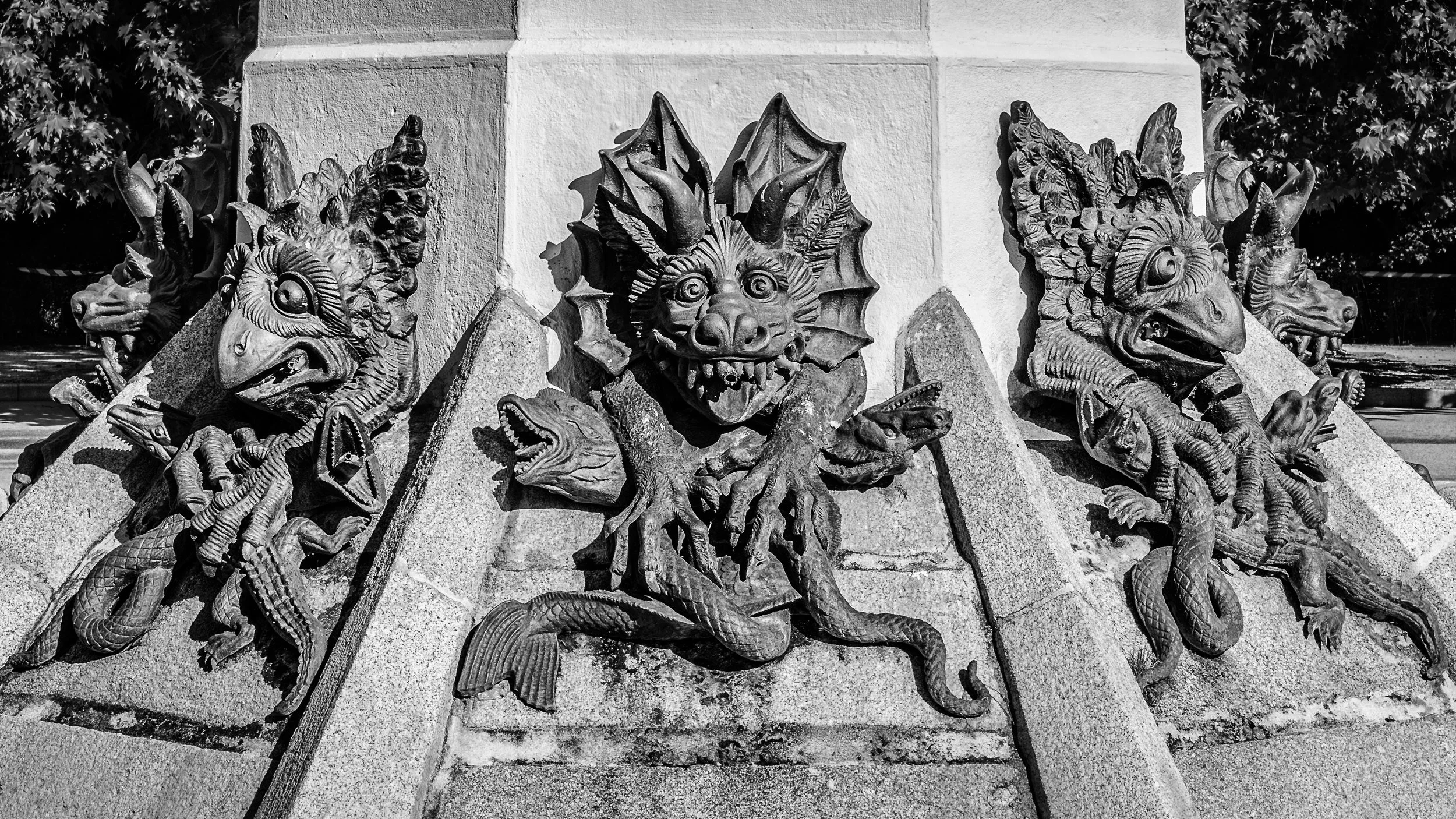 Black and white photo of a decorative stone sculpture featuring mythological creatures and a central dragon face, mounted on a wall or monument with a tree visible in the background.