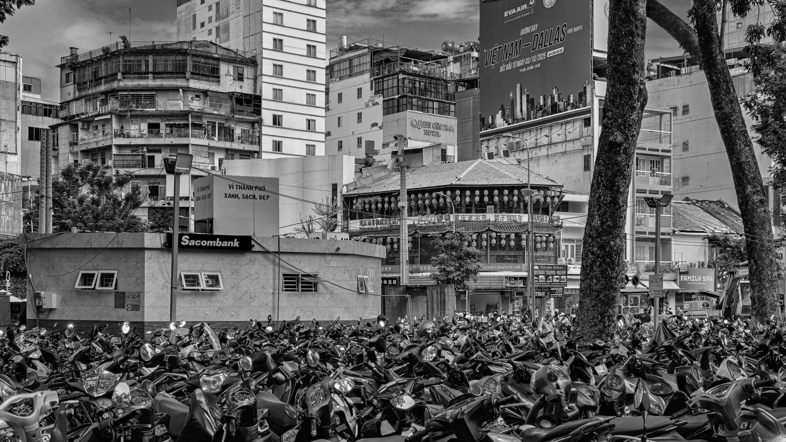 Cityscape with multiple high-rise buildings, a billboard advertising Vietnam to Dallas, and a crowded parking lot filled with motorcycles in black and white.