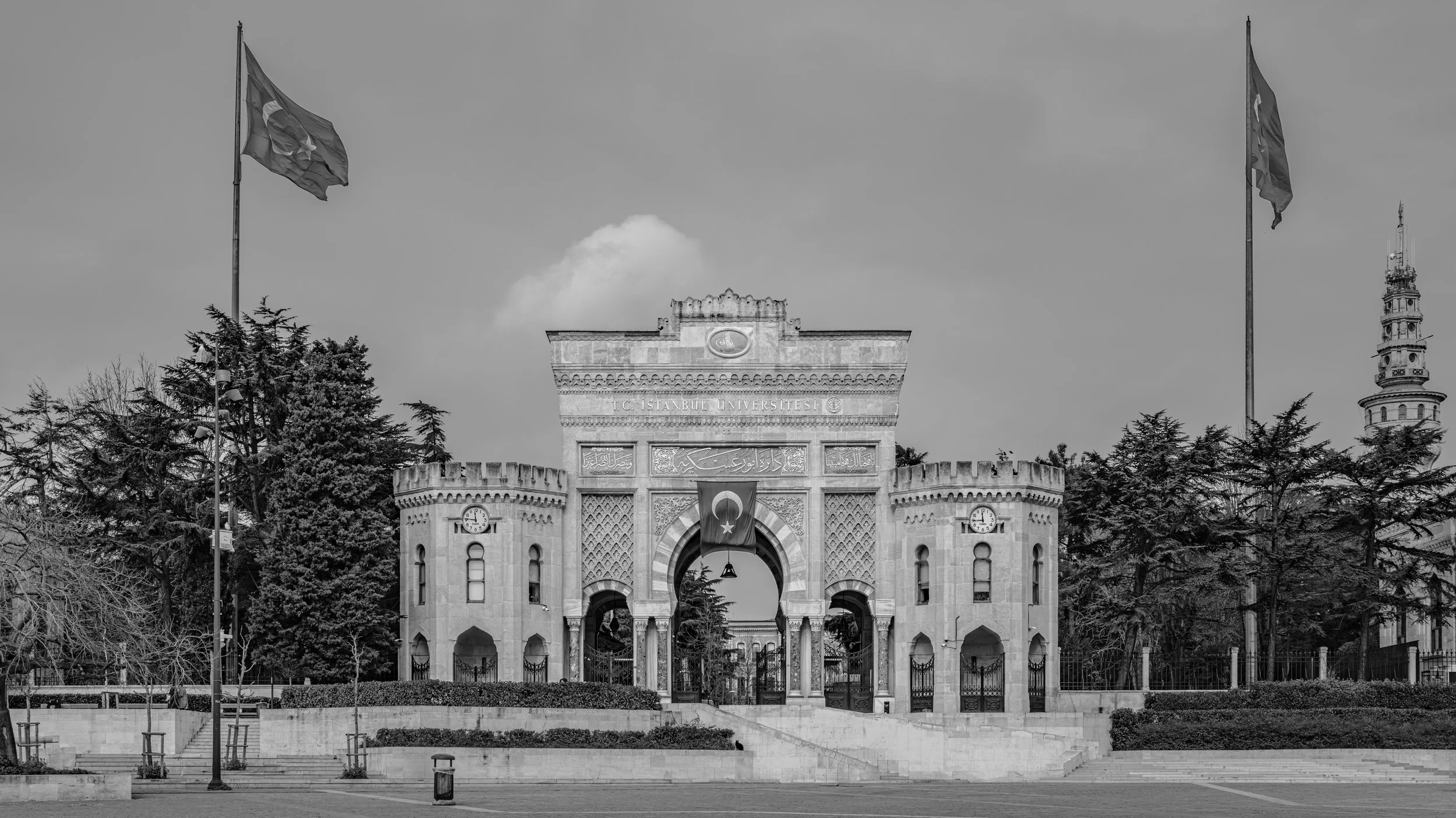 Black and white photo of a historic entrance gate to a university, with two Turkish flags on tall flagpoles, surrounded by trees and a clock on the gate, with a tower visible in the background.