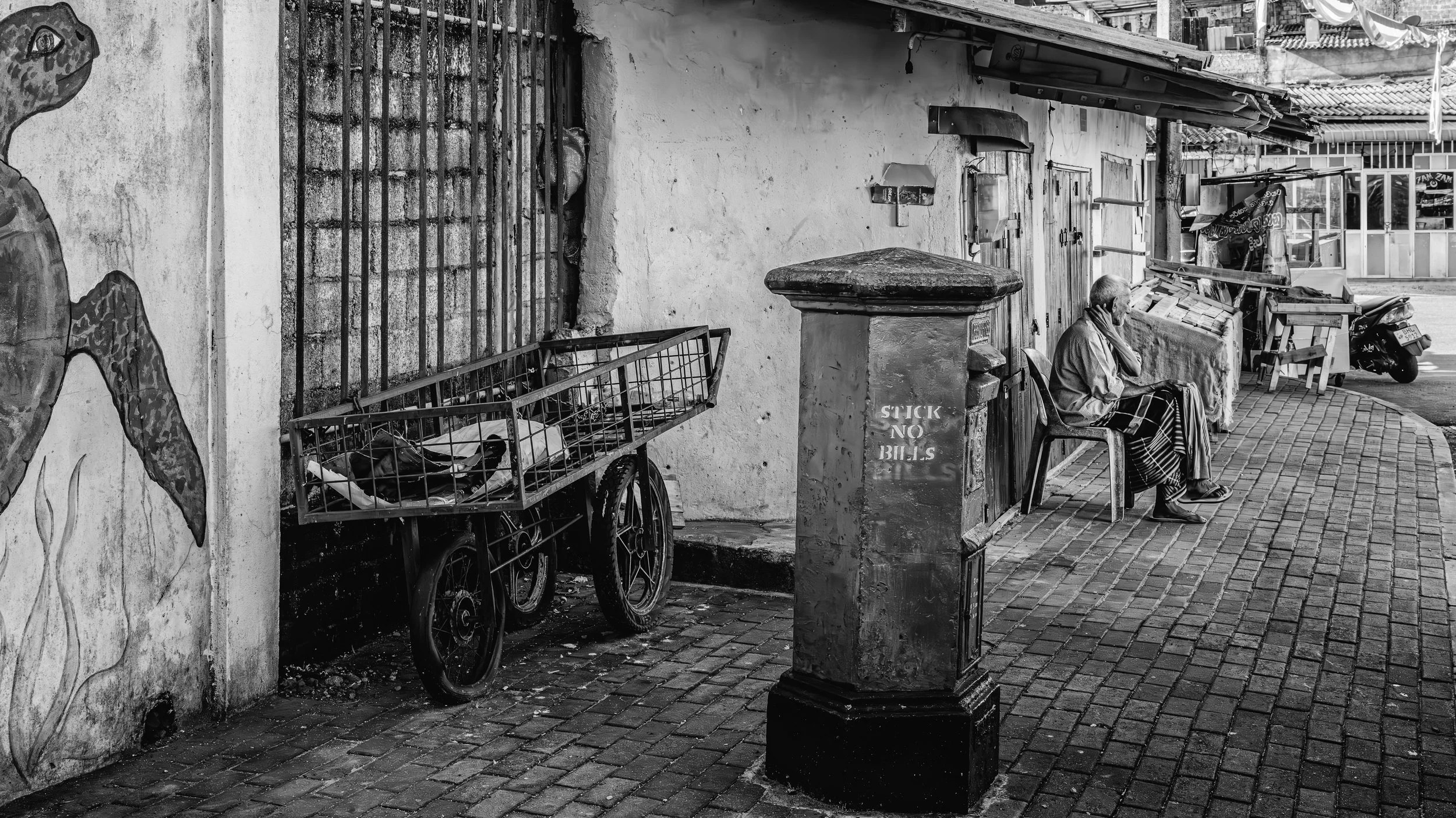A black and white photograph showing an elderly person sitting on a chair on a sidewalk, talking on a phone. There is a cart with items beside them, a wall with a mural, and a street scene with a scooter and shop in the background.