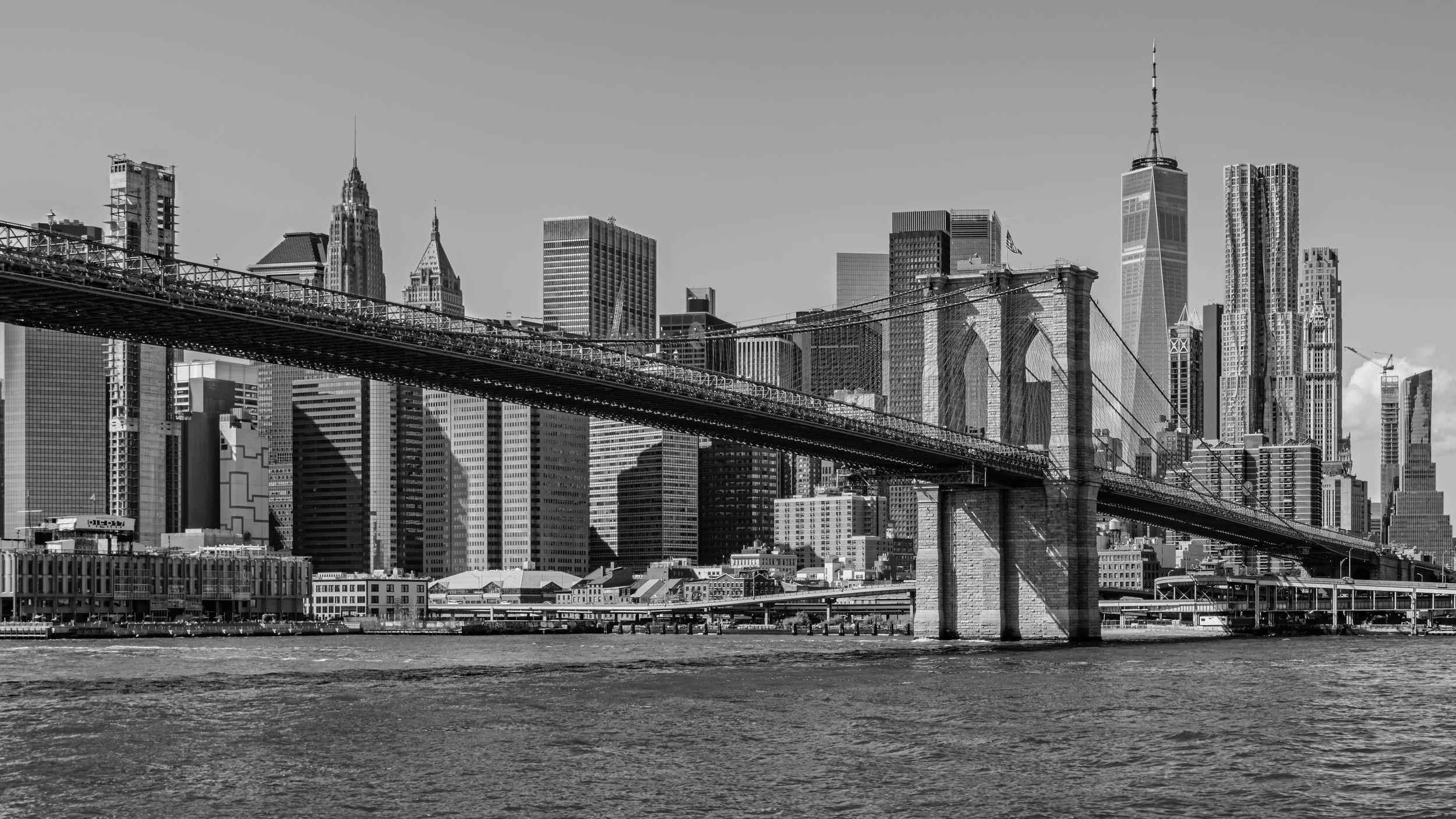 Black and white photo of the New York City skyline viewed from across the river, featuring the Brooklyn Bridge in the foreground and tall skyscrapers including One World Trade Center. Manhattan, New York, USA.