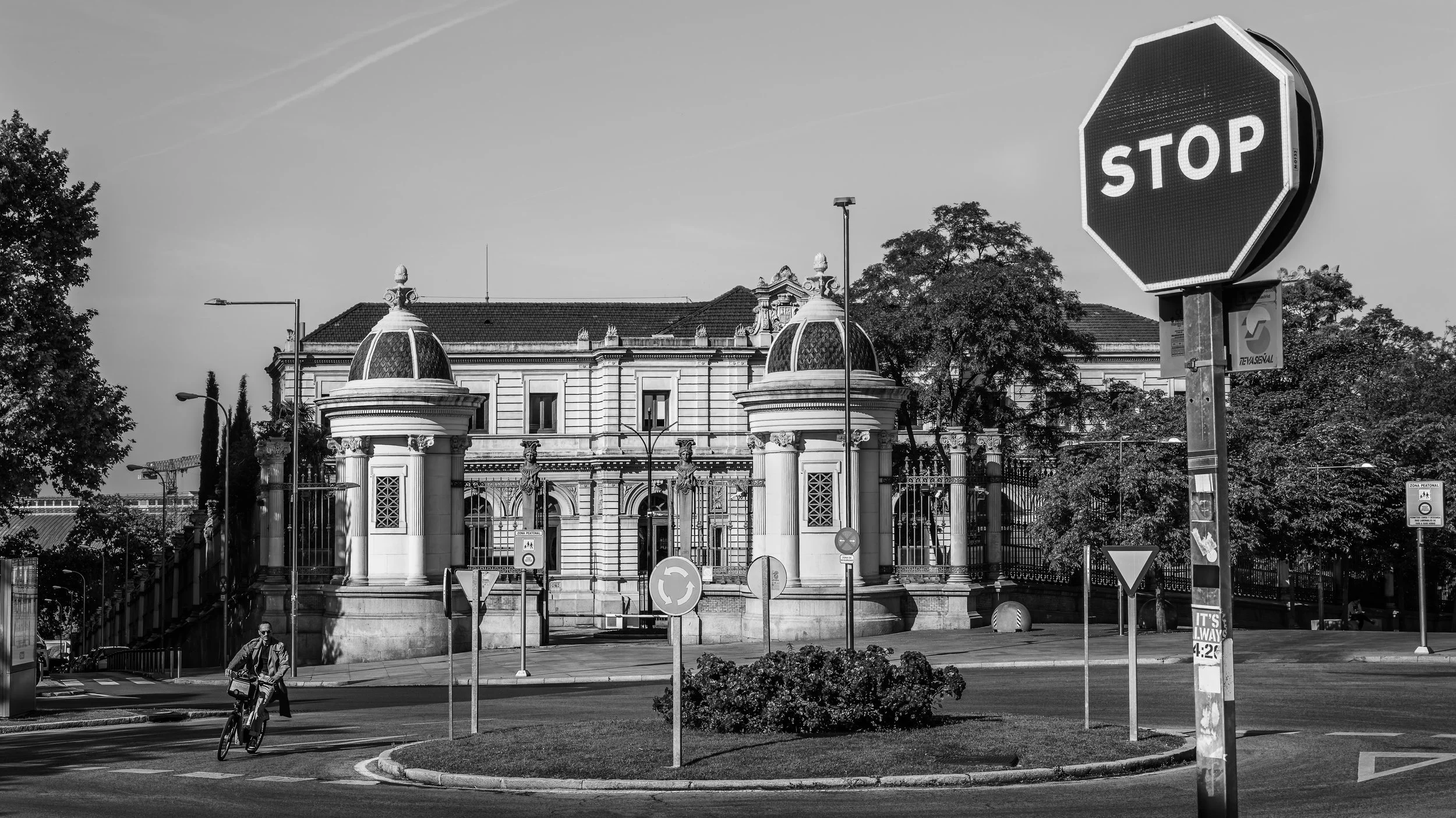 A black-and-white photo of a historic building with ornate architectural details, surrounded by trees, traffic signs, and streetlights. Prominent stop sign in the foreground.