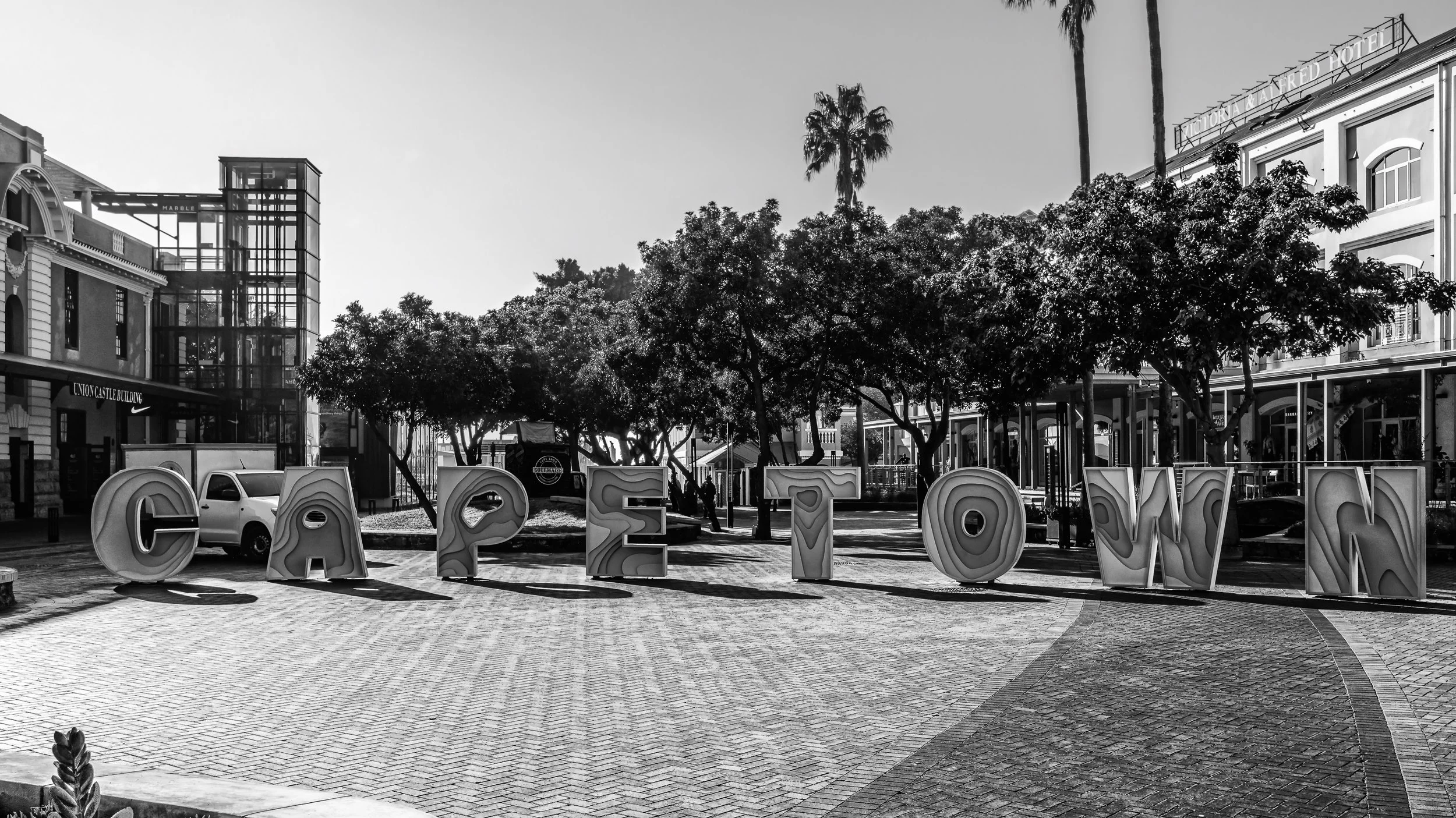 Large decorative letters spelling 'CARMEL' in an outdoor public area with trees and buildings, including a modern glass elevator structure and a hotel, in the background. Cape Town, South Africa.