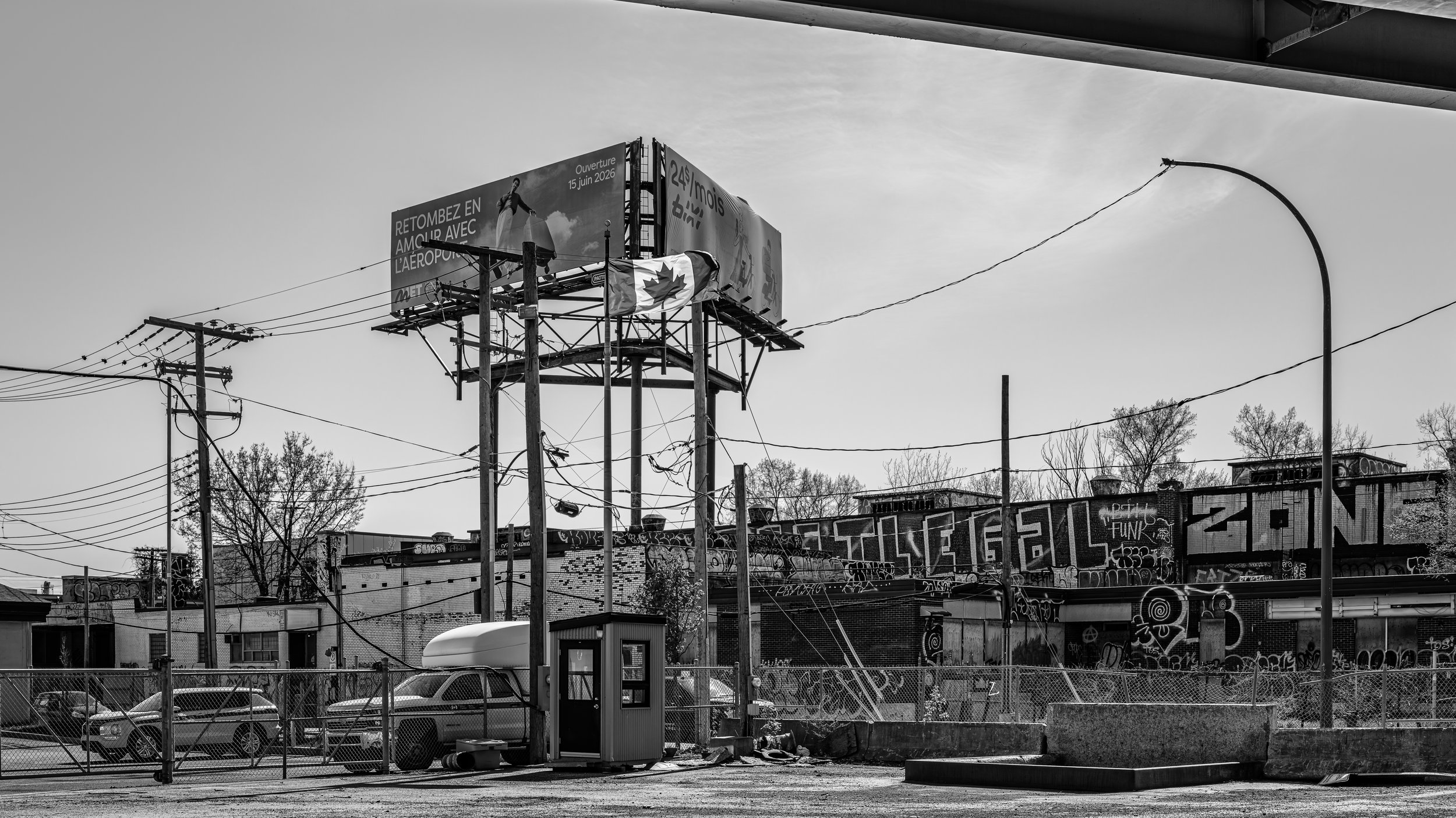 Black and white photo of an urban scene with electric poles and wires, graffiti-covered buildings, and a billboard in the background showing a man and a Canadian flag.