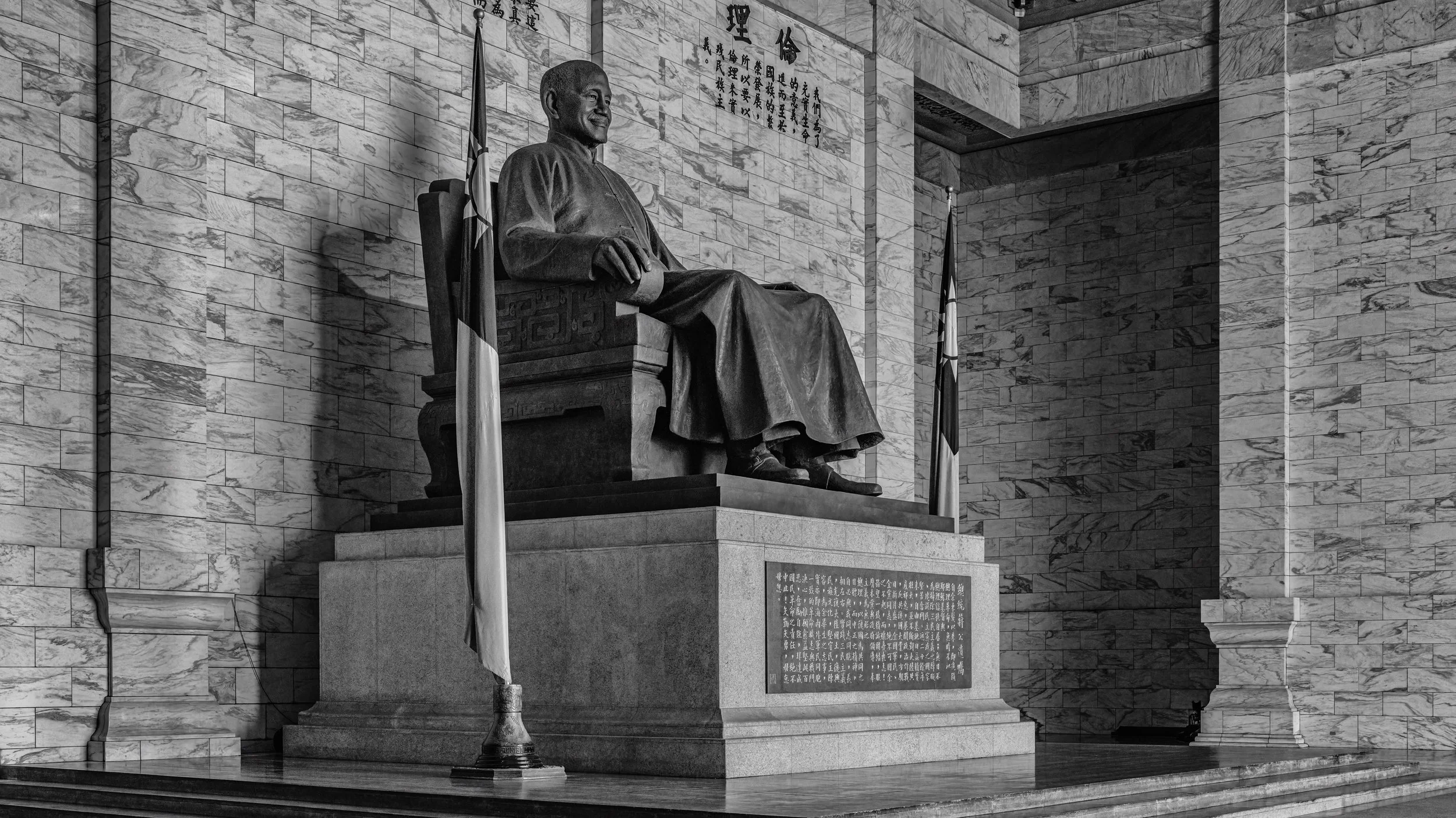 A large bronze statue of Sun Yat-sen seated in a chair, with Chinese characters and flags behind and to the side of the statue, set against a marble wall in a government or historical building.