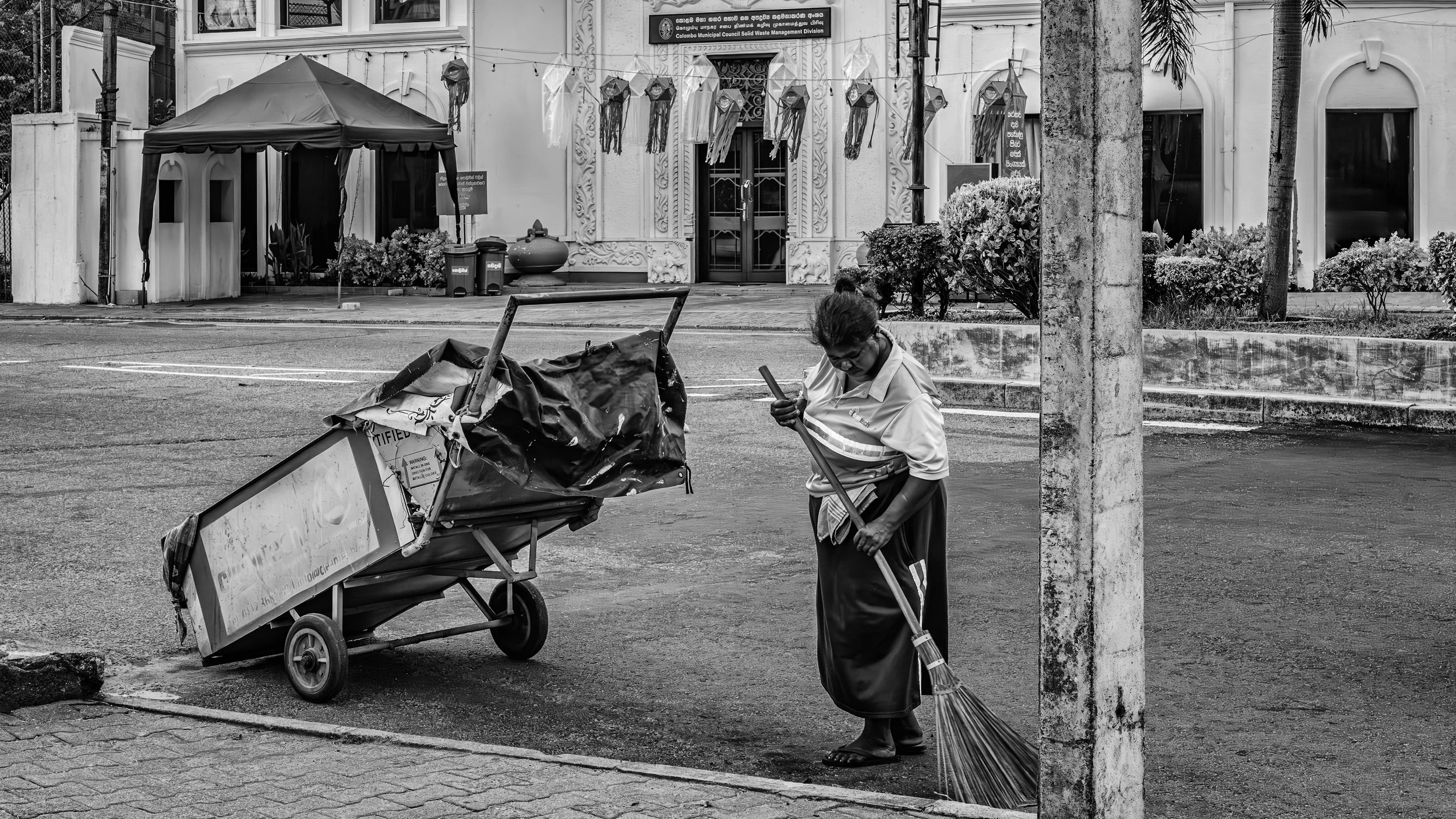A person sweeping the street with a broom near a wheelbarrow, in front of a building with ornate doors and signage, with decorations hanging above.