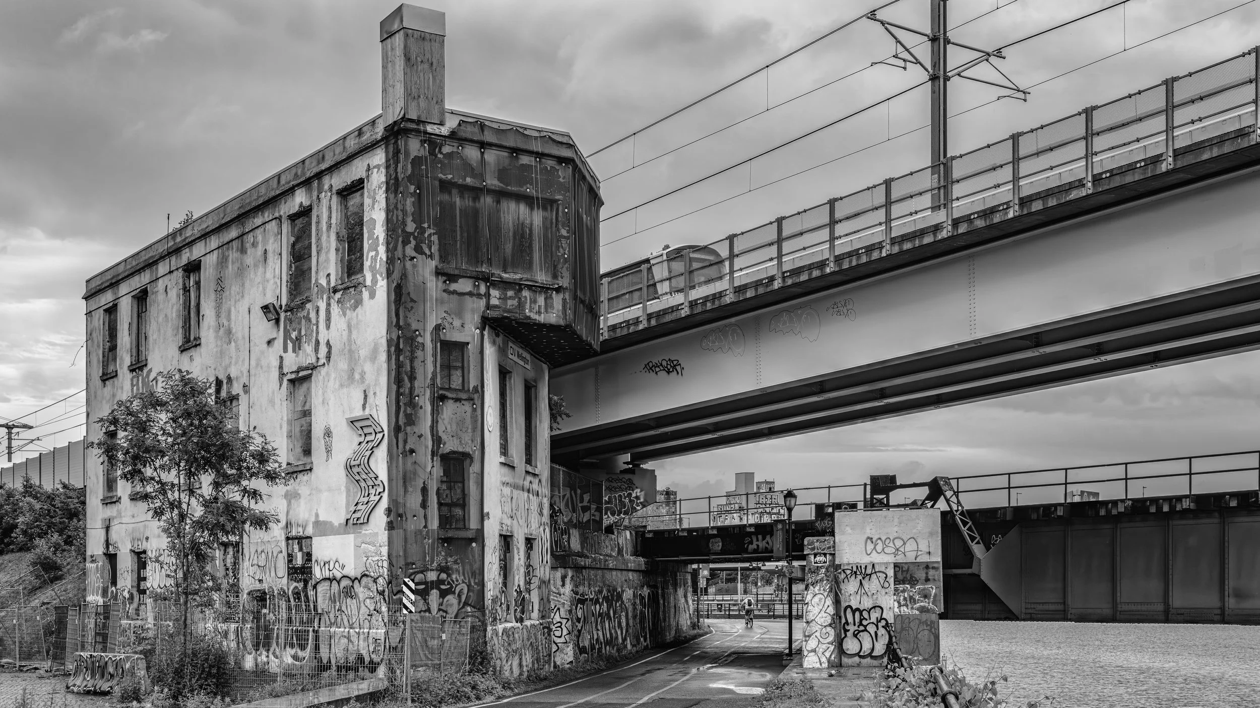 Black and white photograph of an urban scene with an old, weathered building covered in graffiti, next to a modern bridge with train tracks overhead, under a cloudy sky.