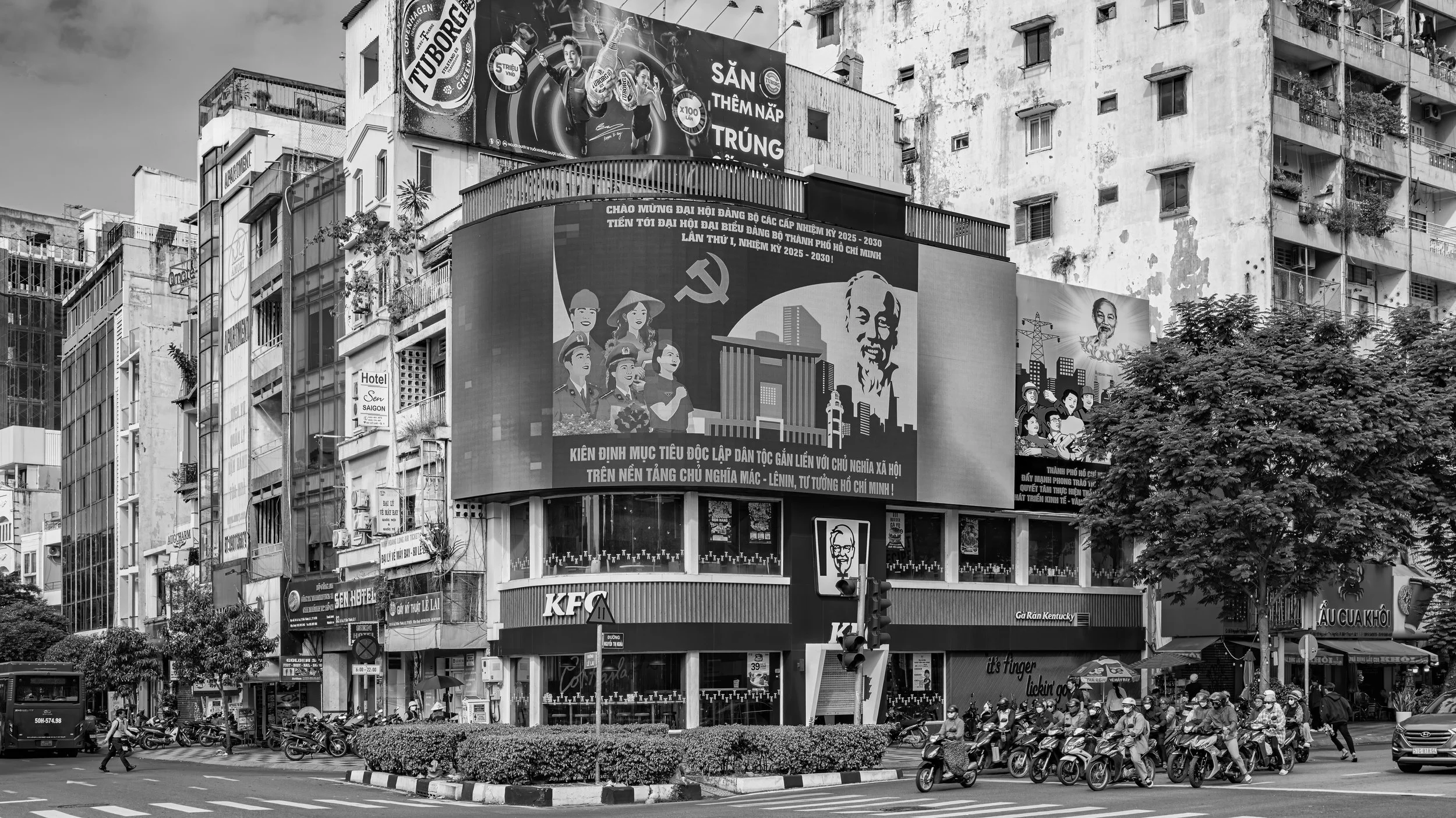 A city street scene with high-rise buildings, billboards, and advertisements. One prominent billboard shows artwork of soldiers and a person with text in Vietnamese. There are numerous motorbikes and people on the street, along with a traffic light. 