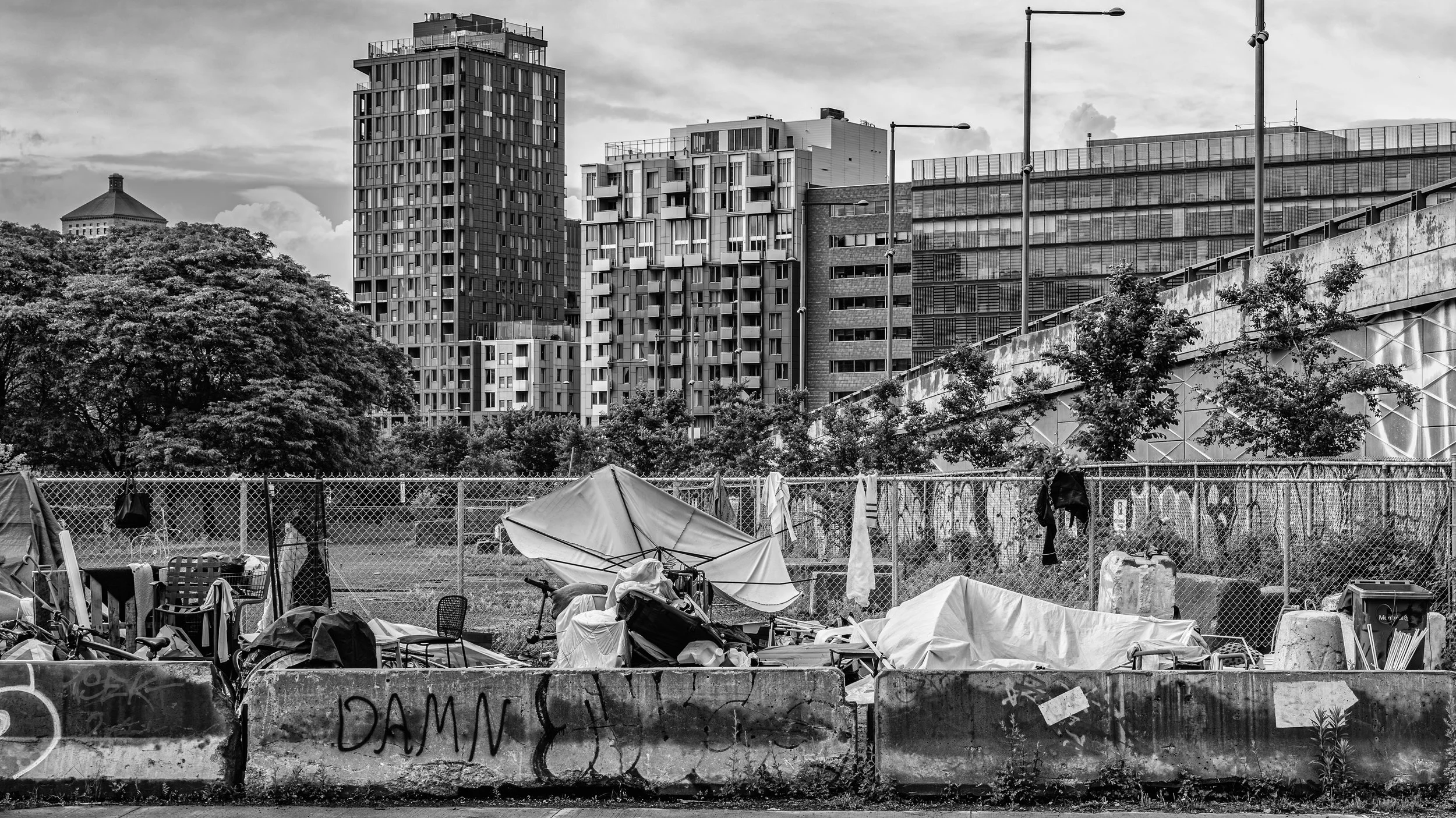 A black and white photo of a cityscape with tall modern buildings, trees, and clouds in the background. In the foreground, there is a fenced area with various tents, chairs, bicycles, and scattered items.