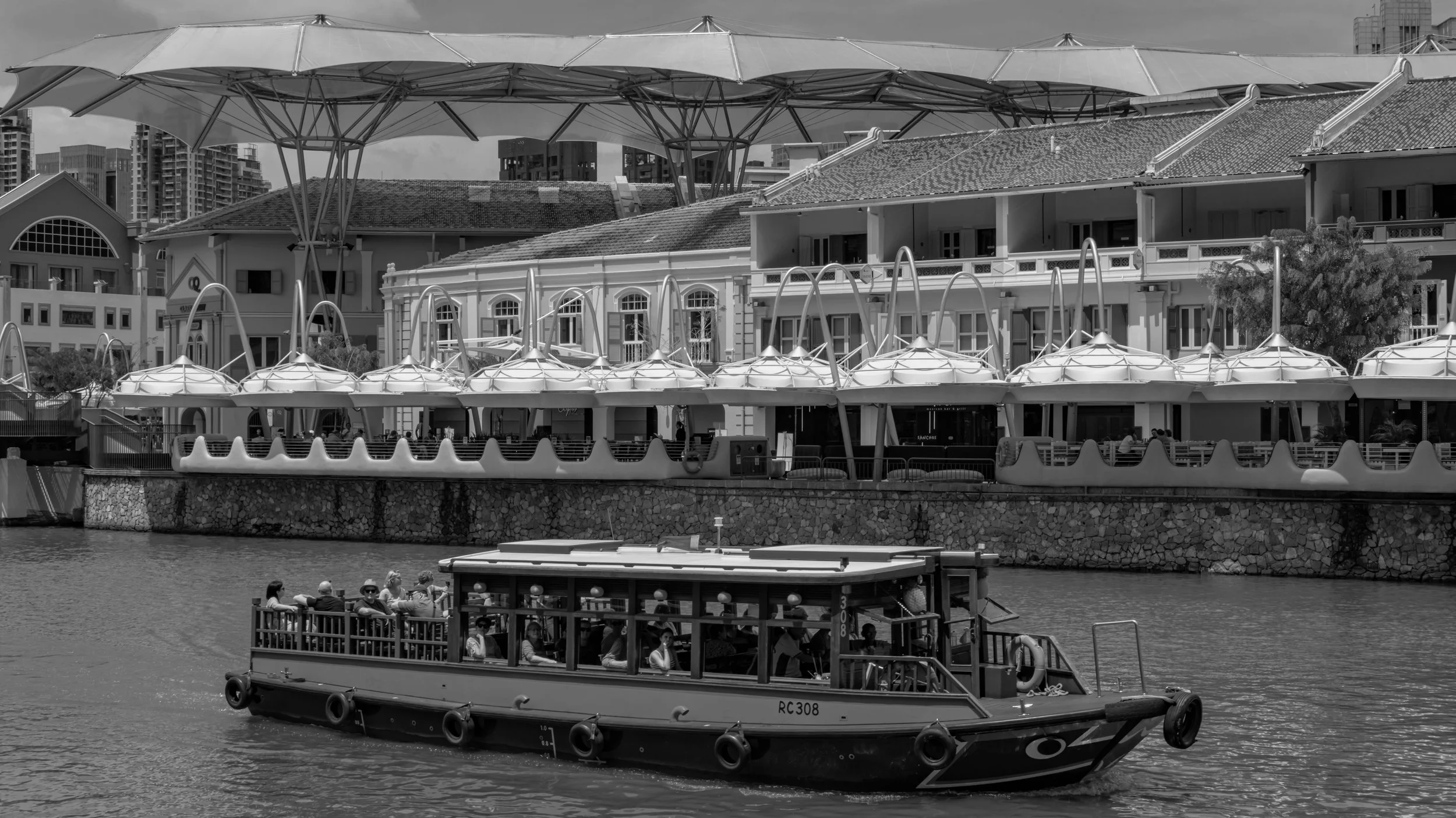 A boat with passengers on a body of water in front of a multi-story building with a large umbrella-like structure on the rooftop. The scene is in black and white.