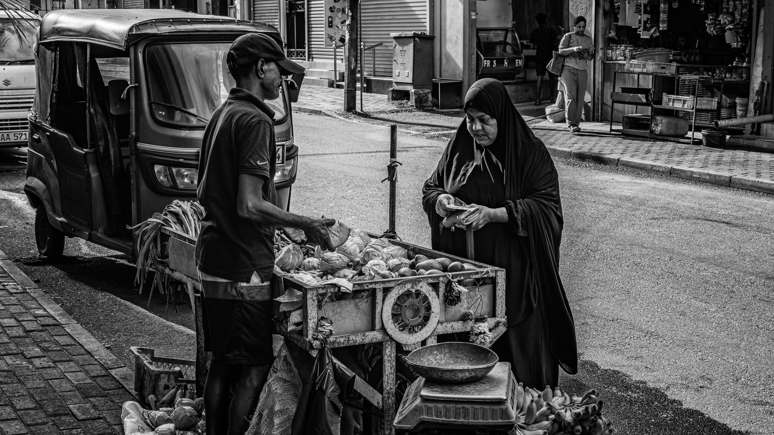A woman in a hijab buying fruit from a street vendor on a city sidewalk, with other pedestrians and storefronts in the background.