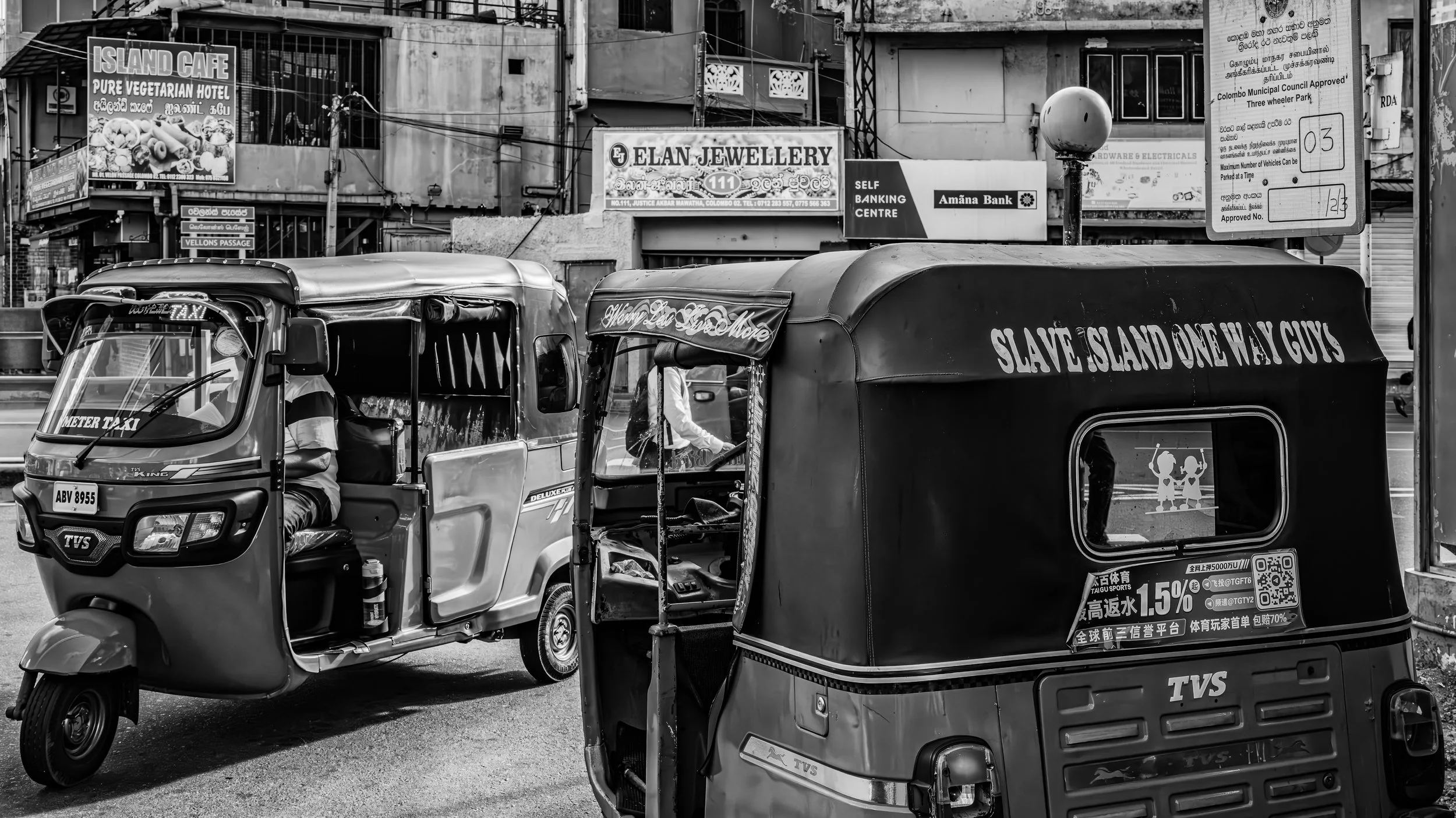 A street scene with auto rickshaws and various signs in an urban area, including an ad for an island cafe, a jewelry store, a bank, and a parking sign, all in black and white.