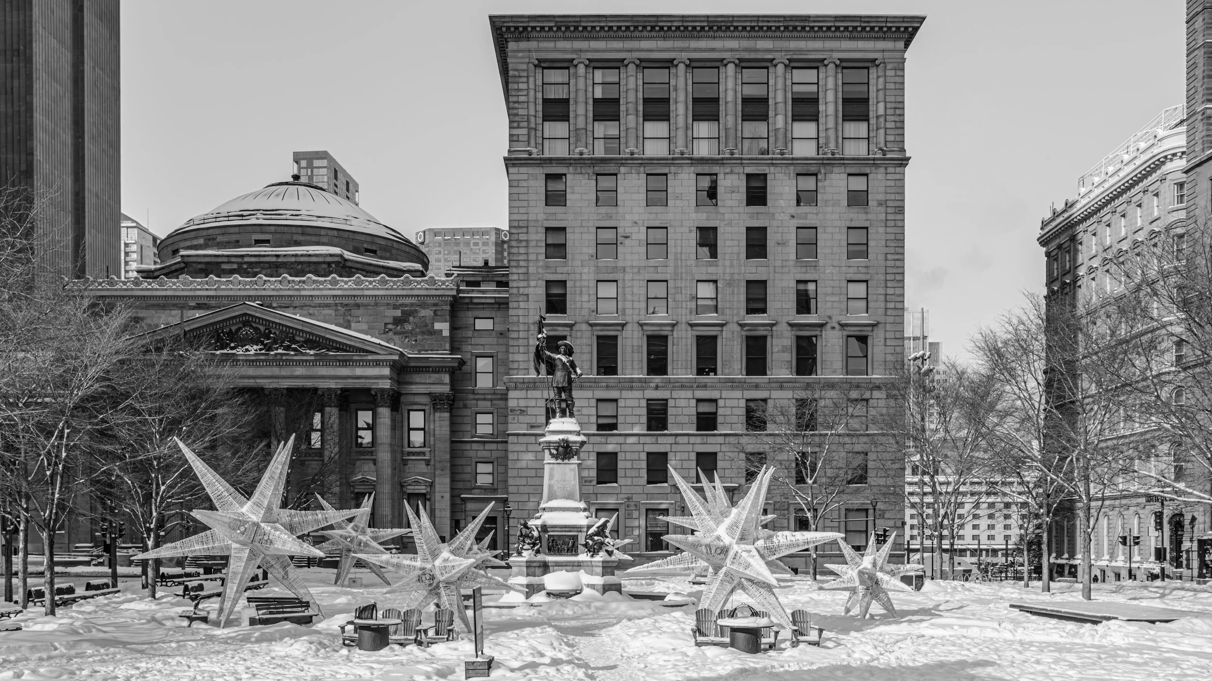 Black and white photo of a city park with snow, decorated for Christmas, featuring a statue on a pedestal, star-shaped ornaments, benches, leafless trees, and tall buildings, including one with ancient architectural style and another modern skyscrape