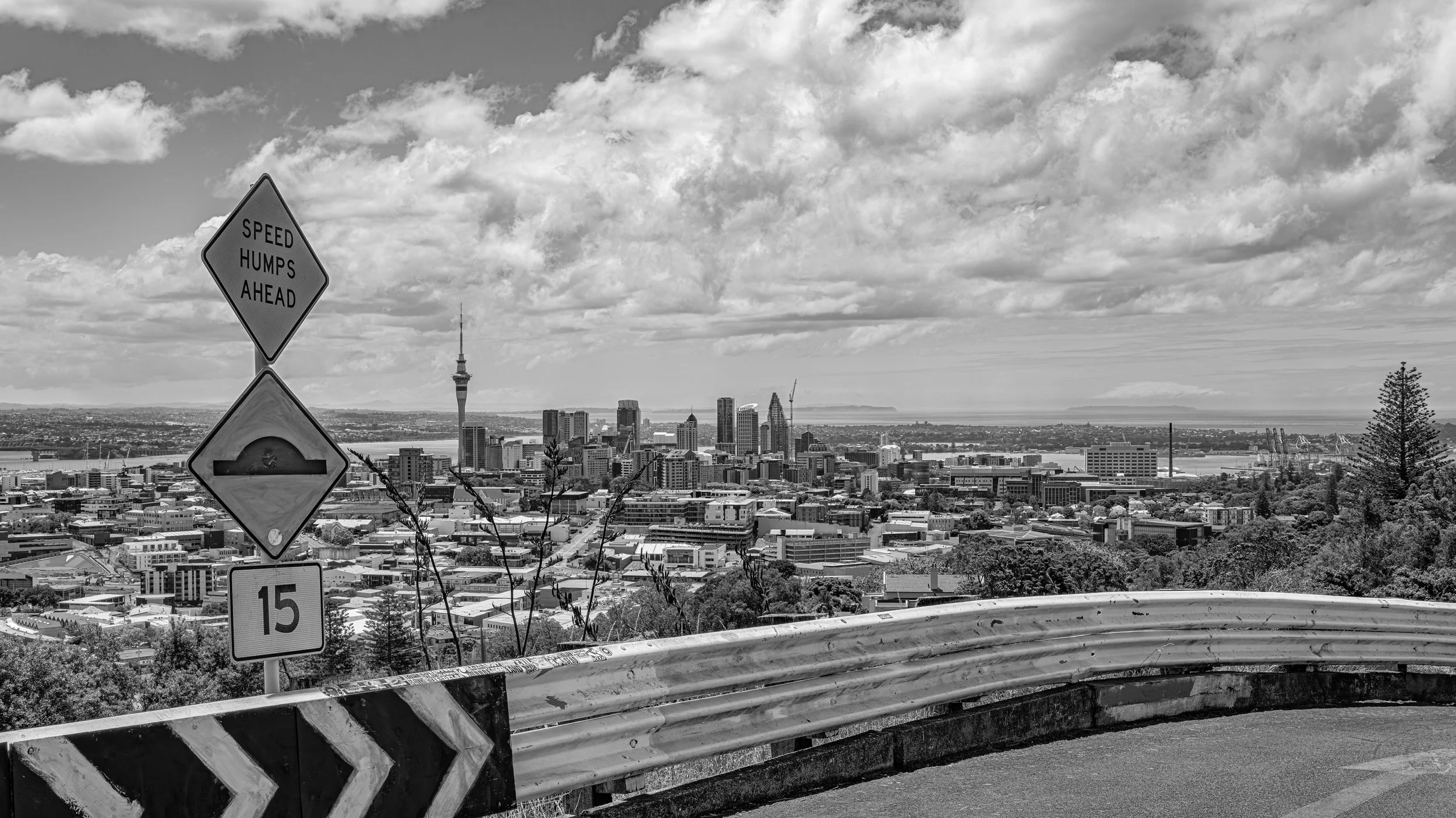 Black and white photo of a city skyline with tall buildings and a tower in the distance, viewed from a hillside with traffic signs and a guardrail in the foreground. The sky is filled with clouds.
