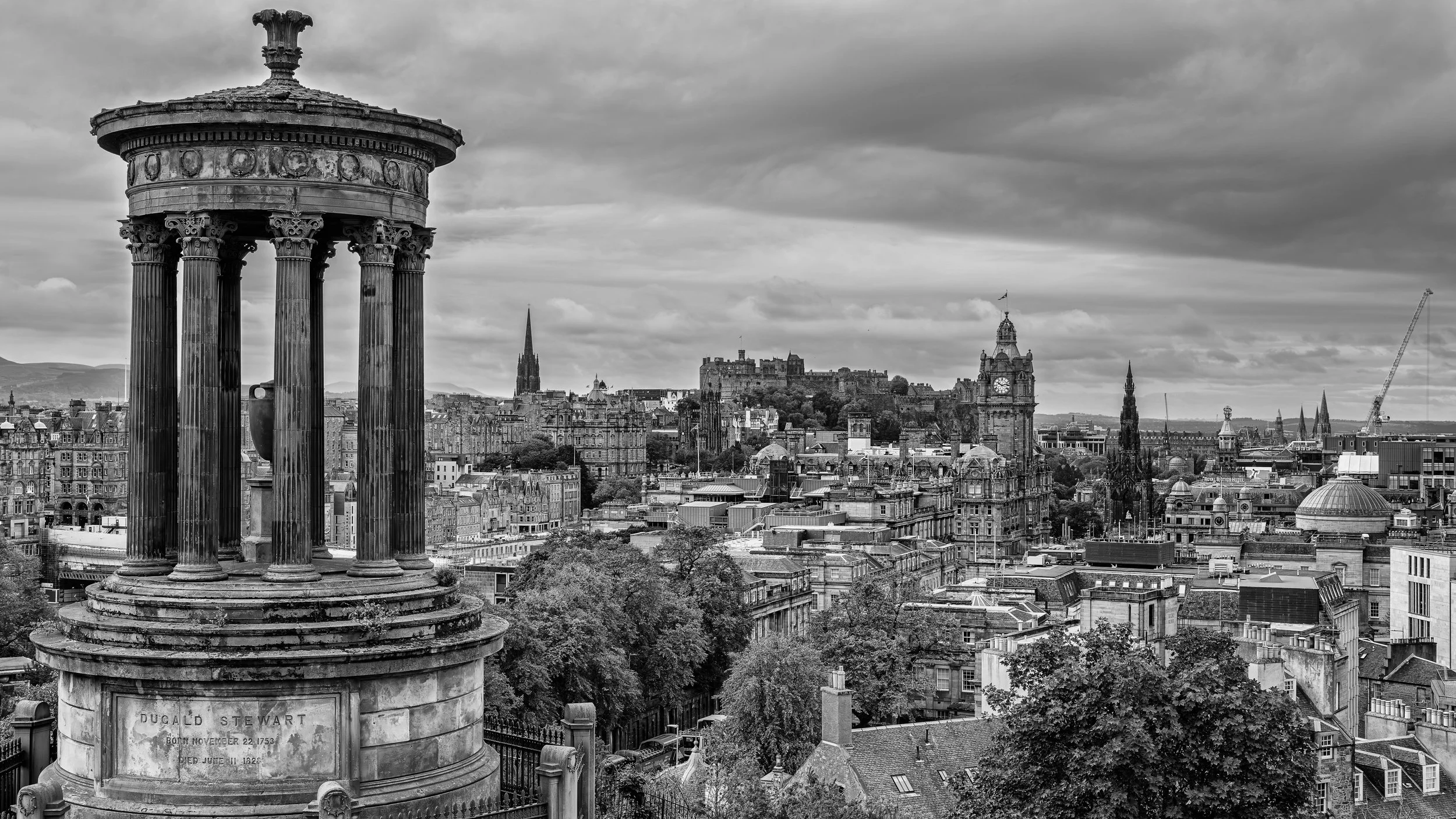 Black and white photo of Edinburgh cityscape with Dugald Stewart monument in foreground, historic buildings, churches, and a castle on a hill under cloudy sky.