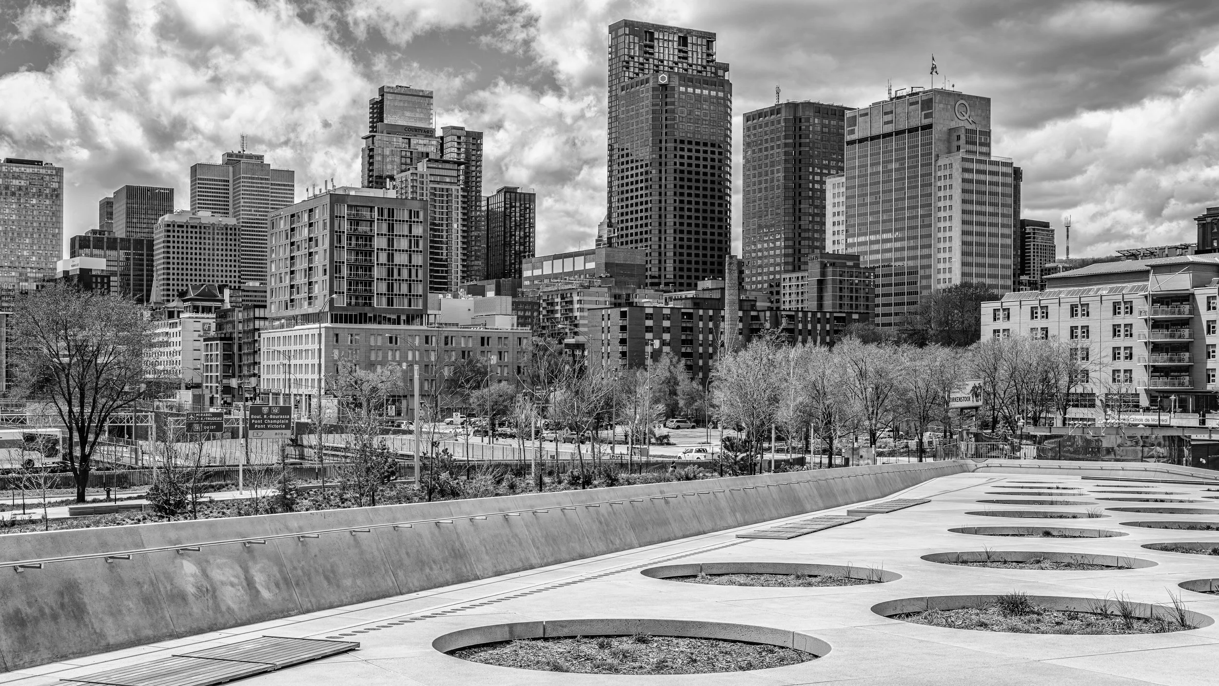 A city skyline with tall buildings and skyscrapers under a cloudy sky, with a foreground of leafless trees and a modern, circular architectural feature.