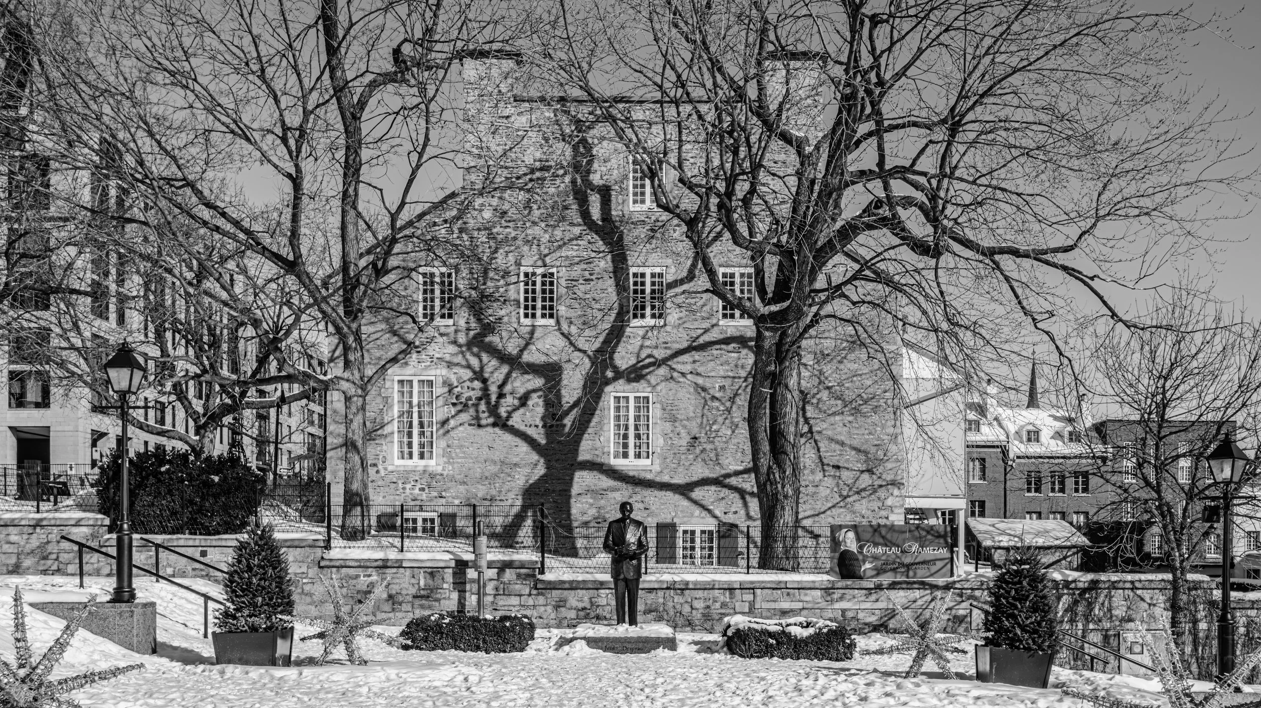 A black and white photo of a historic stone building with several windows, surrounded by leafless trees casting shadows. In the foreground, there is a small park area with snow on the ground, a few small bushes, and a statue of a person.