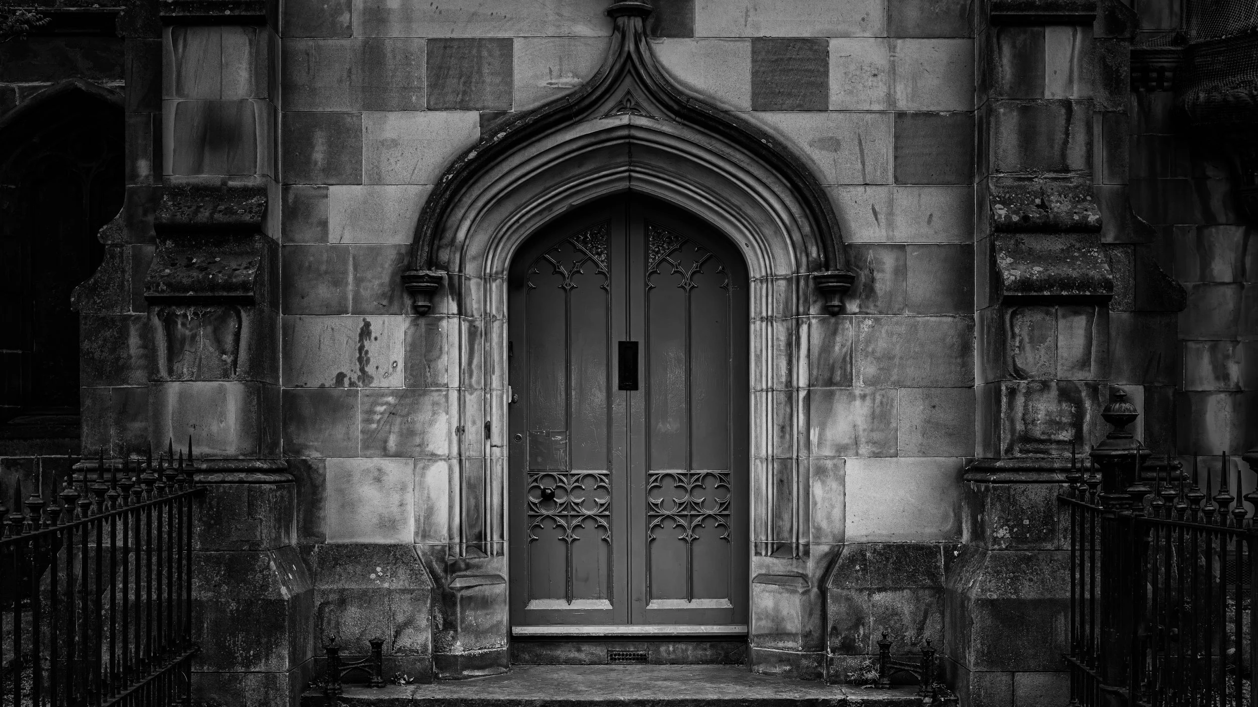 Black and white photo of a stone building entrance with an arched door and decorative ironwork. The doorway is framed by detailed stone carvings and flanked by stone columns, with a small gate in front.