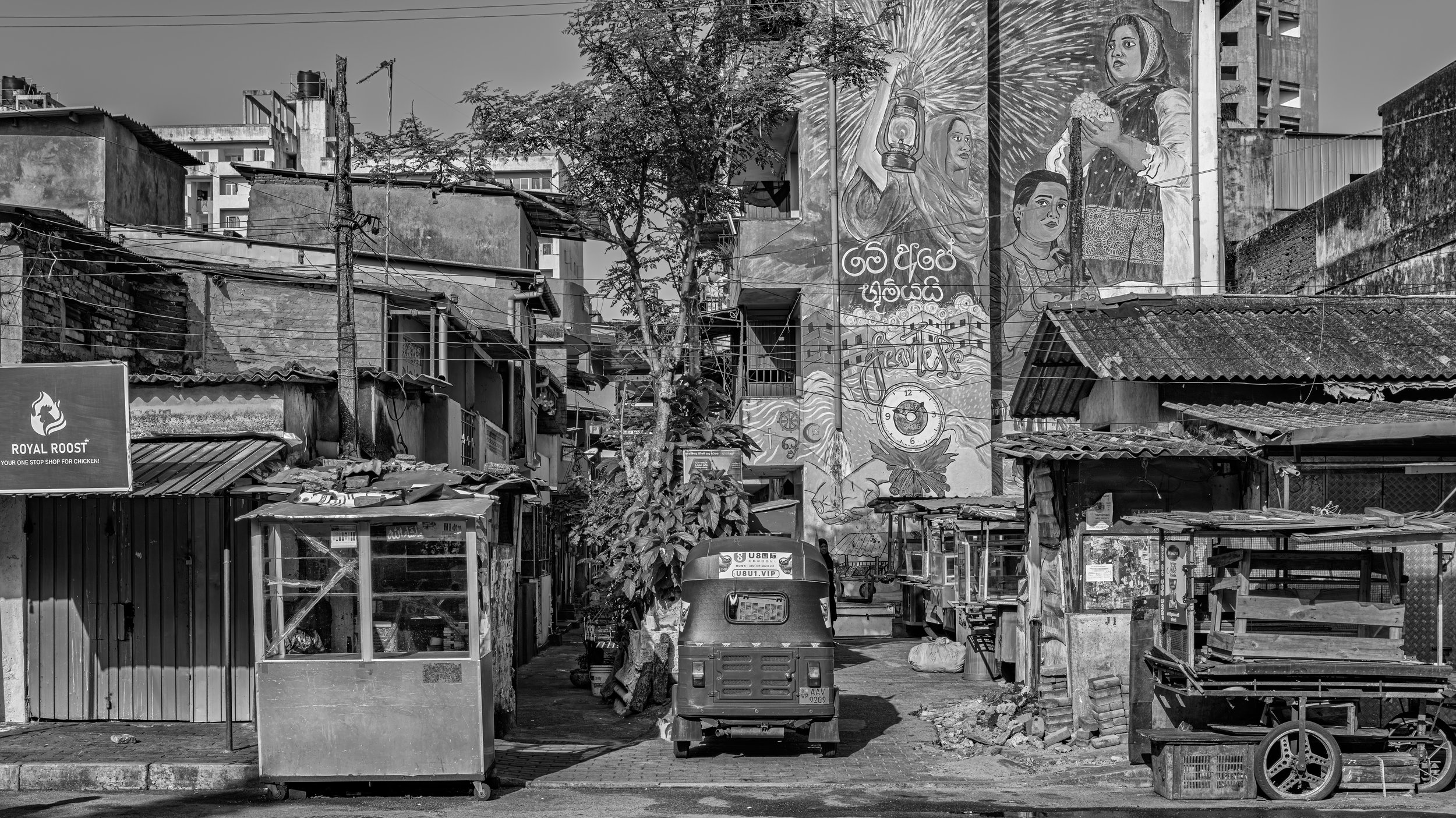 A black and white photo of an urban scene with graffiti murals on a large wall featuring women holding lanterns, set amidst makeshift structures and buildings, electrical wires, a tree, and a small auto rickshaw in the foreground.