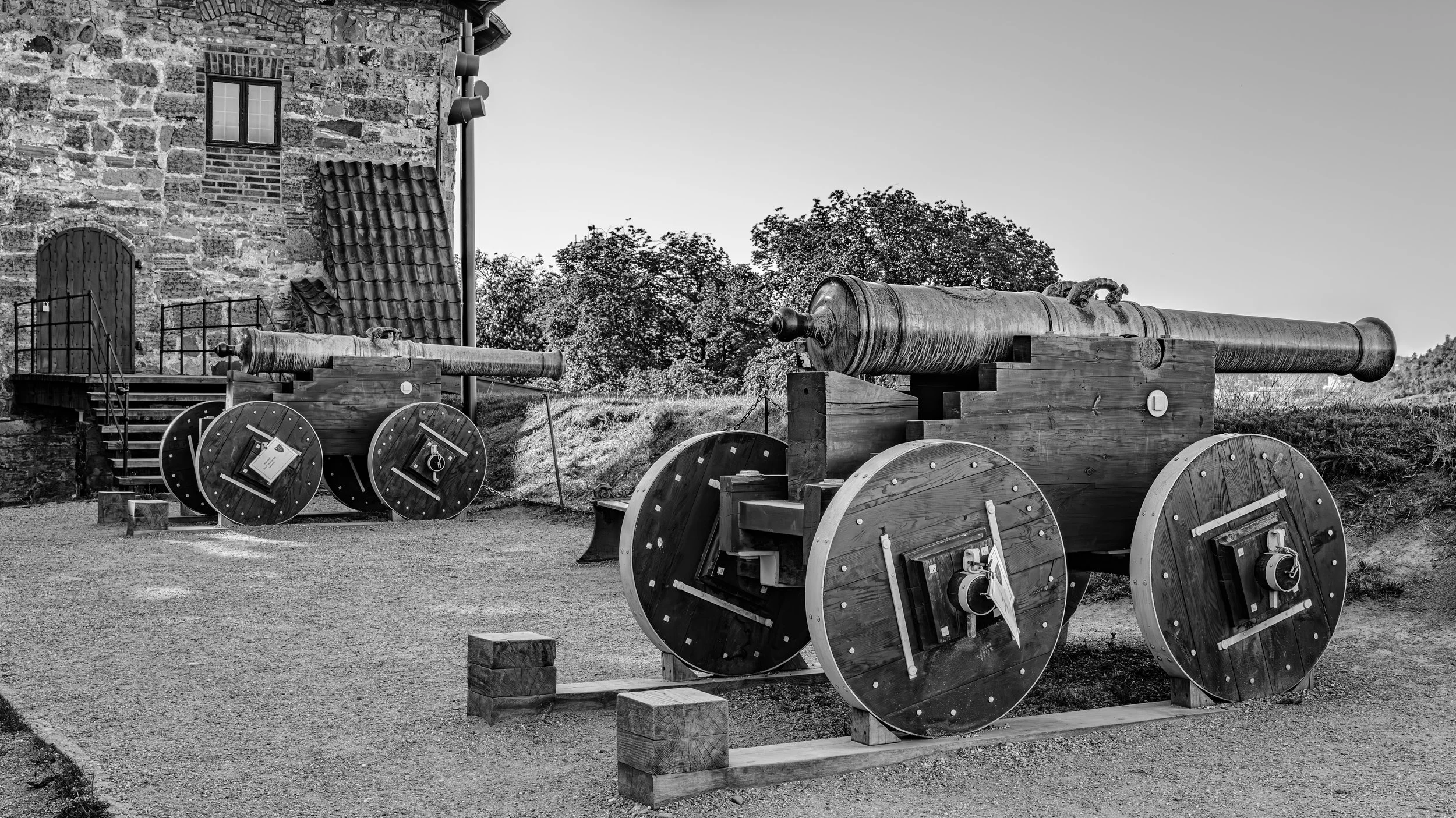 Black and white photograph of old cannons mounted on wooden wheels outside a stone building, with trees in the background.