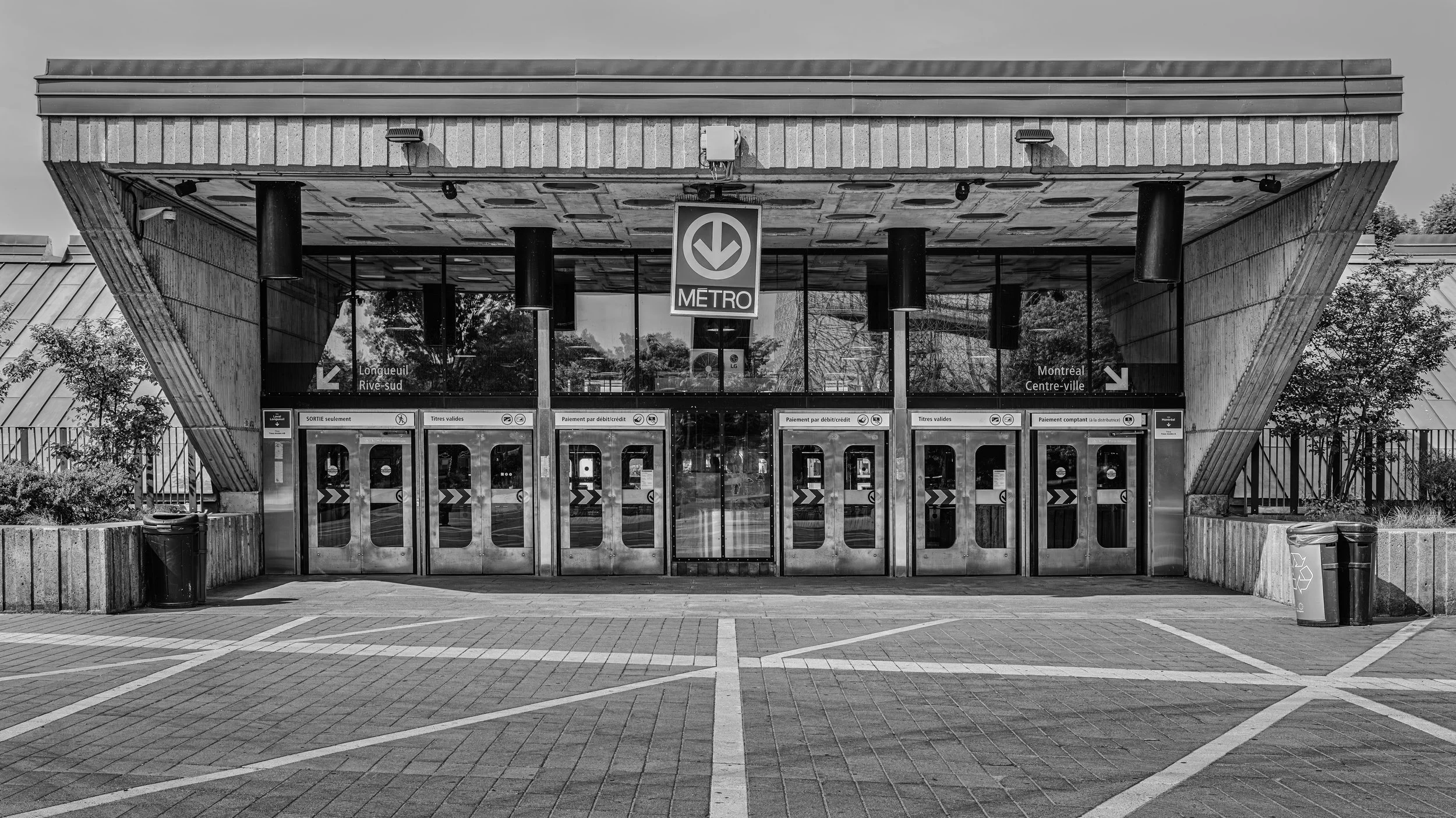 Black and white photo of a metro station entrance with glass doors. The station is escalator-accessible with signs indicating directions to Longueuil, Rive-sud, Montreal, and Centre-ville. The station features a modern architectural style with concre