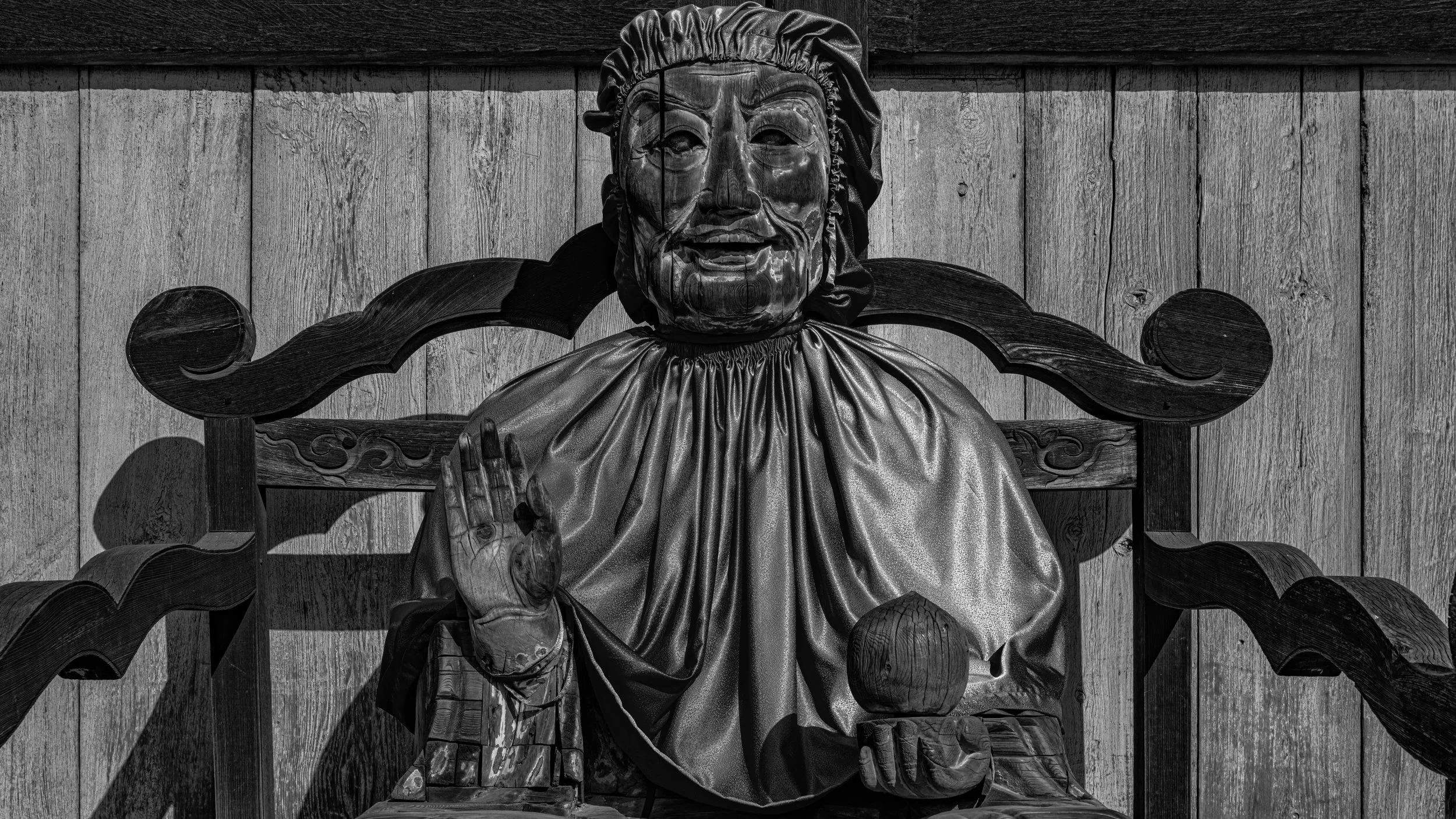 Black and white photo of a wooden sculpture of a bearded man sitting on a carved wooden throne, with a wooden wall background.