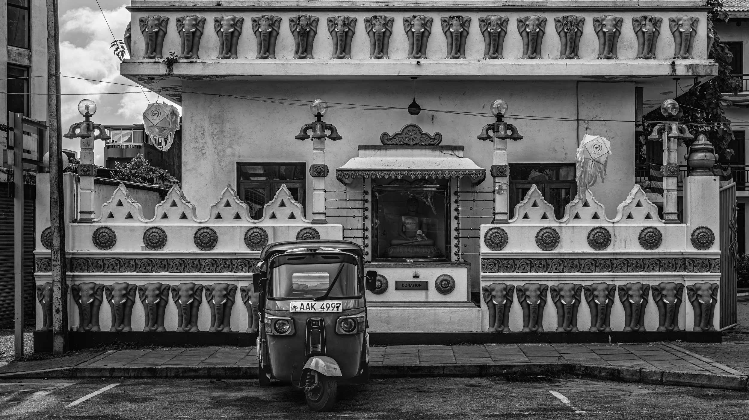 A two-story building with decorative elephant head sculptures along the railing of the balcony and along the fence in front. There are two hanging lanterns, a statue of Buddha behind the glass door, and a small donation box on the porch. A tuk-tuk ve