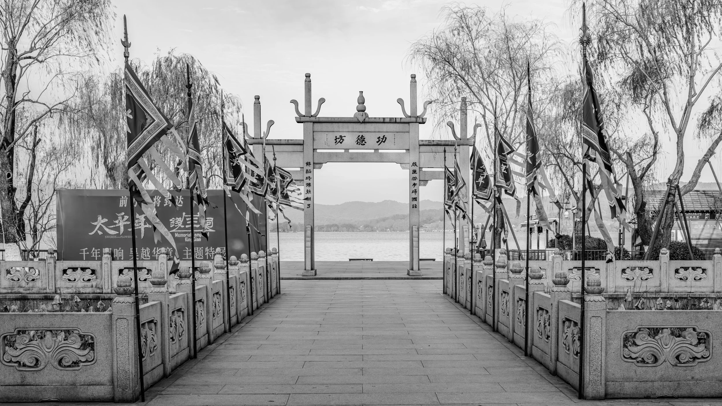 A traditional Chinese stone archway with Chinese characters, flanked by flags and trees, overlooking a body of water with mountains in the background.