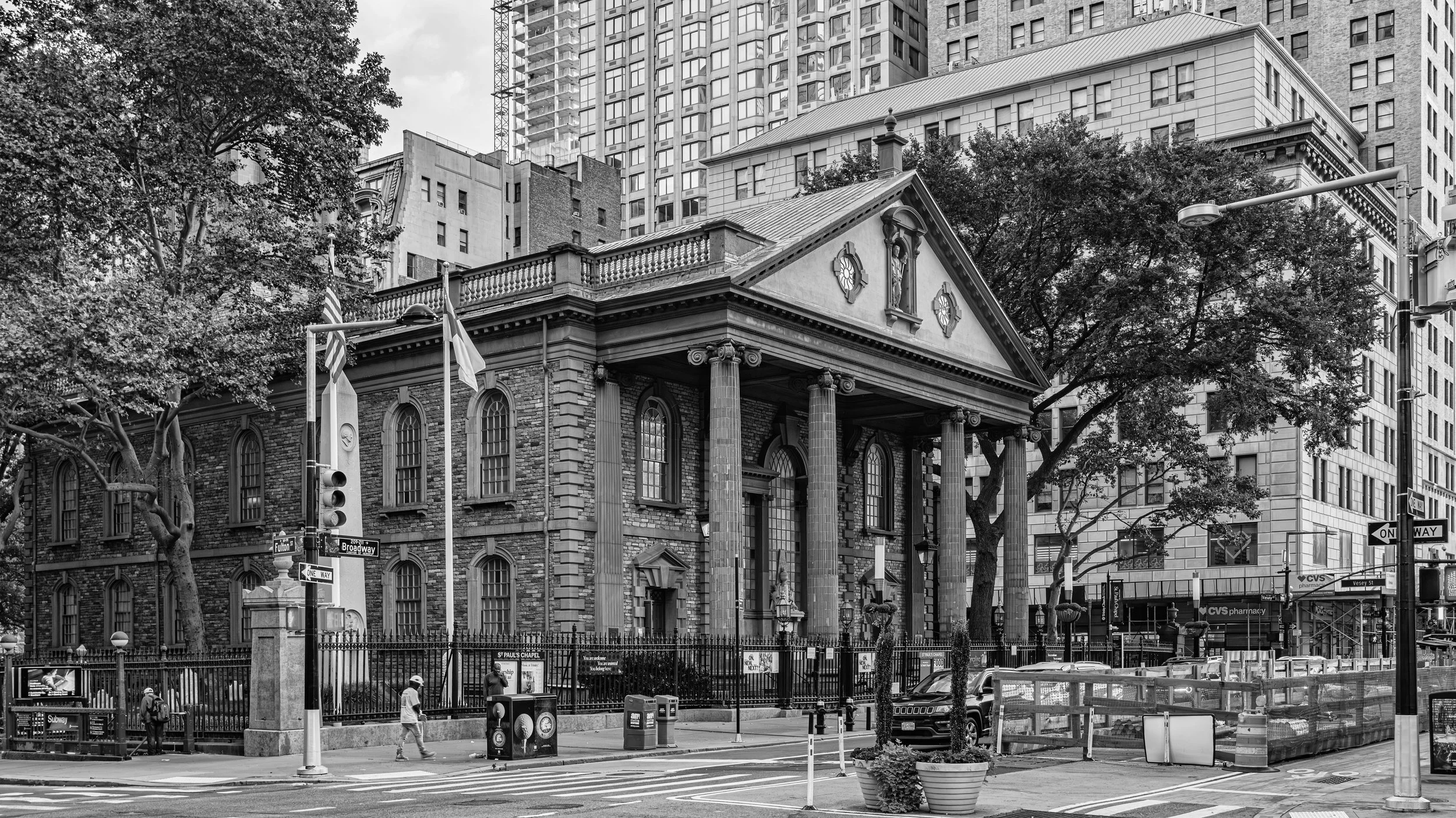 A historic church with tall columns and a pediment, set against modern skyscrapers in an urban environment. There are trees, street signs, and people walking on the sidewalk. Manhattan, New York, USA.