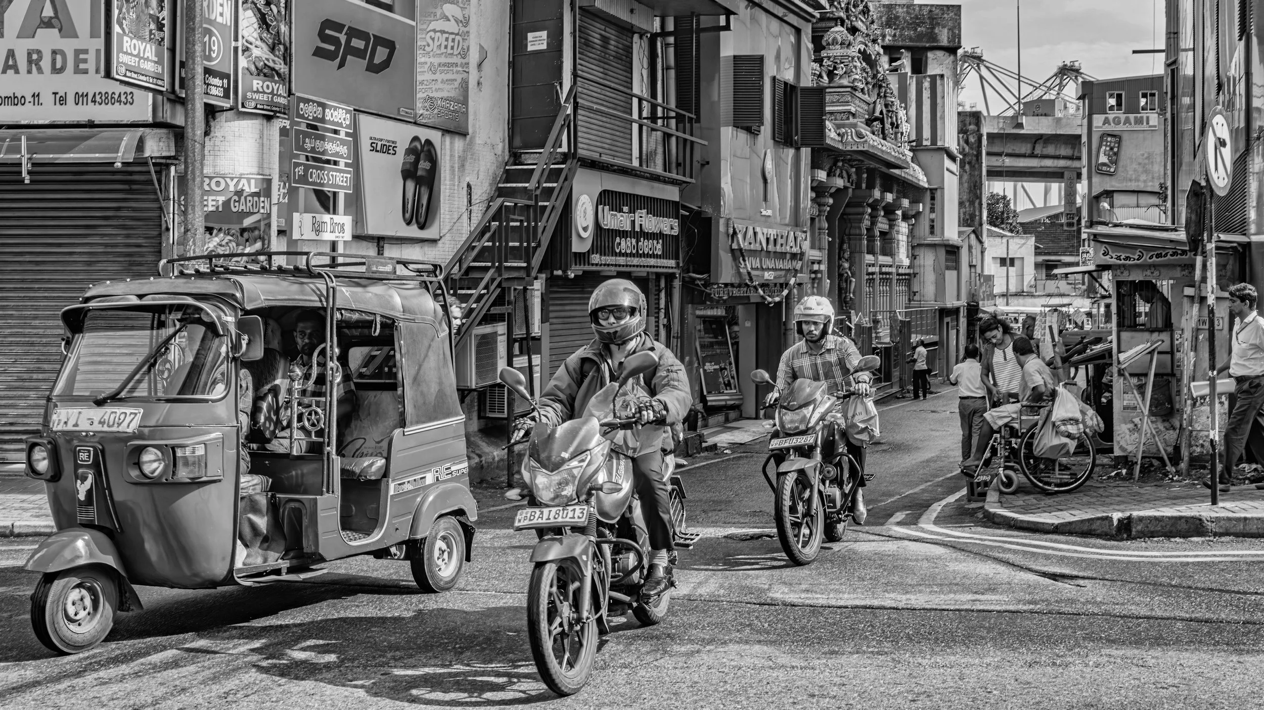 Street scene with motorcycles, auto rickshaw, and people walking and standing near shops in an urban area.