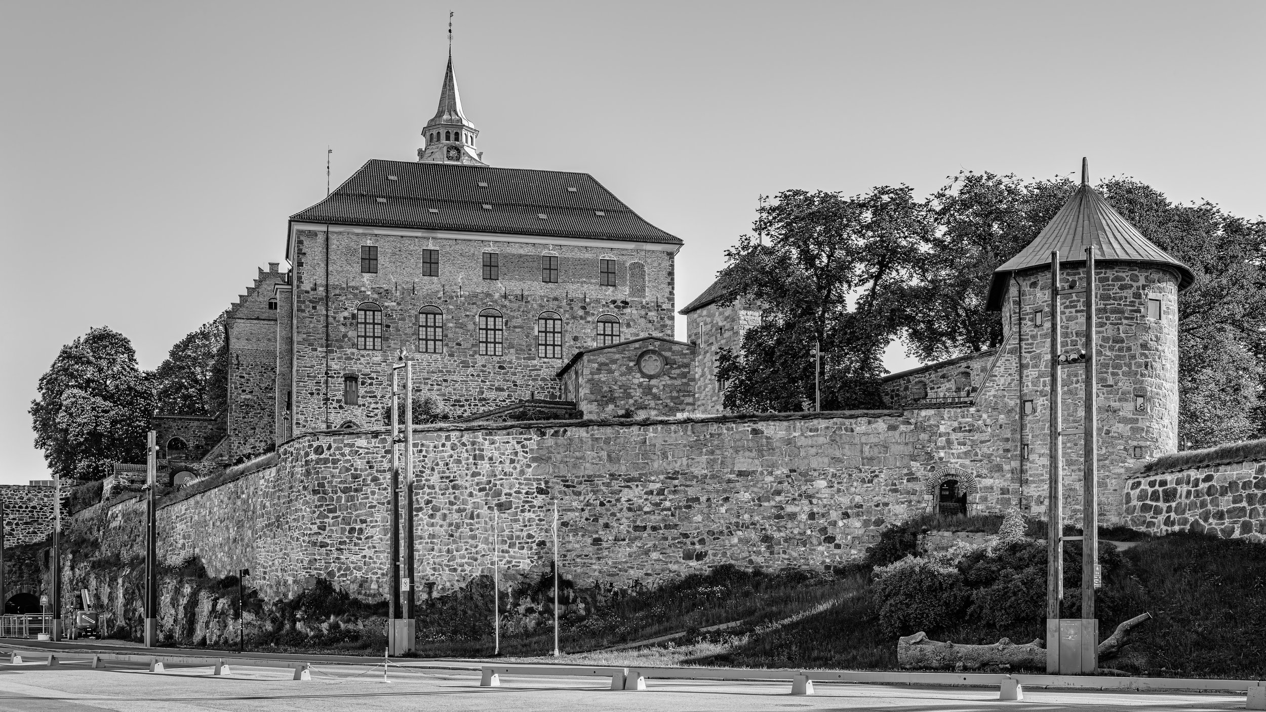 A historic stone castle with a tall, pointed turret, situated on a hill, with large arched windows and a slate roof. There is a stone fortress wall at the base and trees in the background.