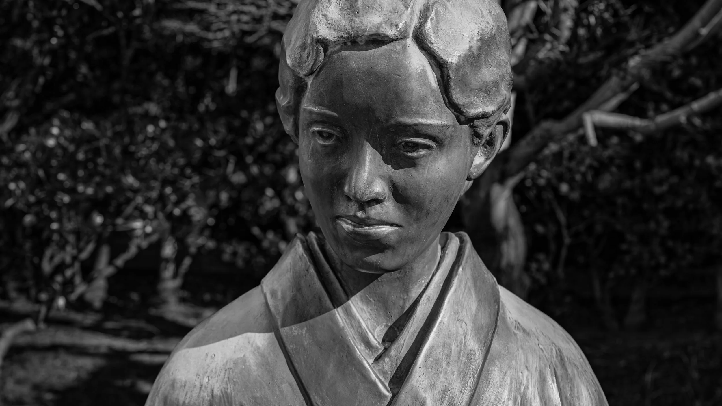 Black and white photo of a sculpture bust of a young boy with short hair, wearing a collared shirt and a jacket, with a serious expression, set against a blurred outdoor background with leaves and branches.