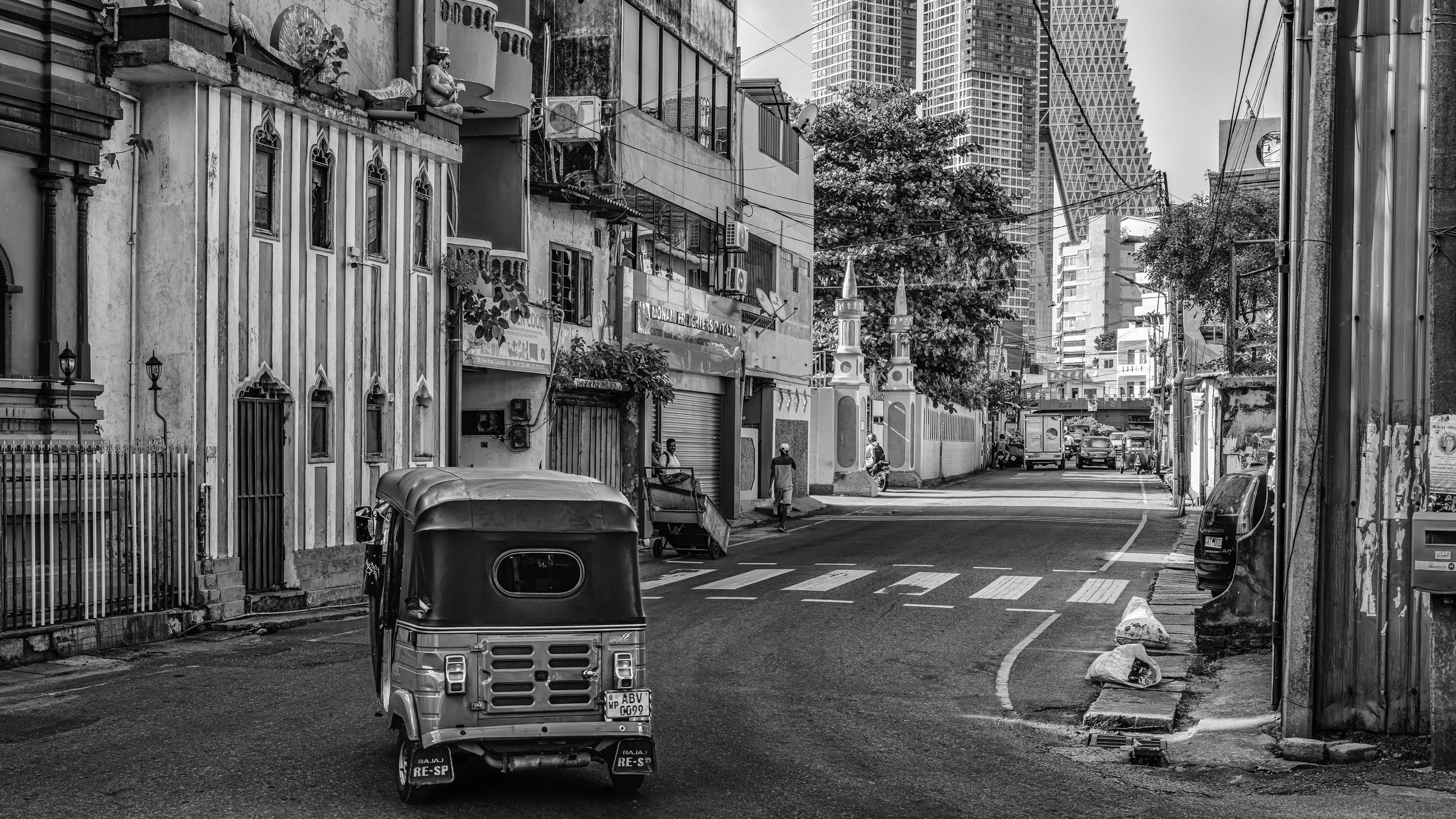 Black and white photo of a narrow city street with buildings, some modern and some older. A small vehicle is in the foreground, along with people walking. Tall skyscrapers are visible in the background.