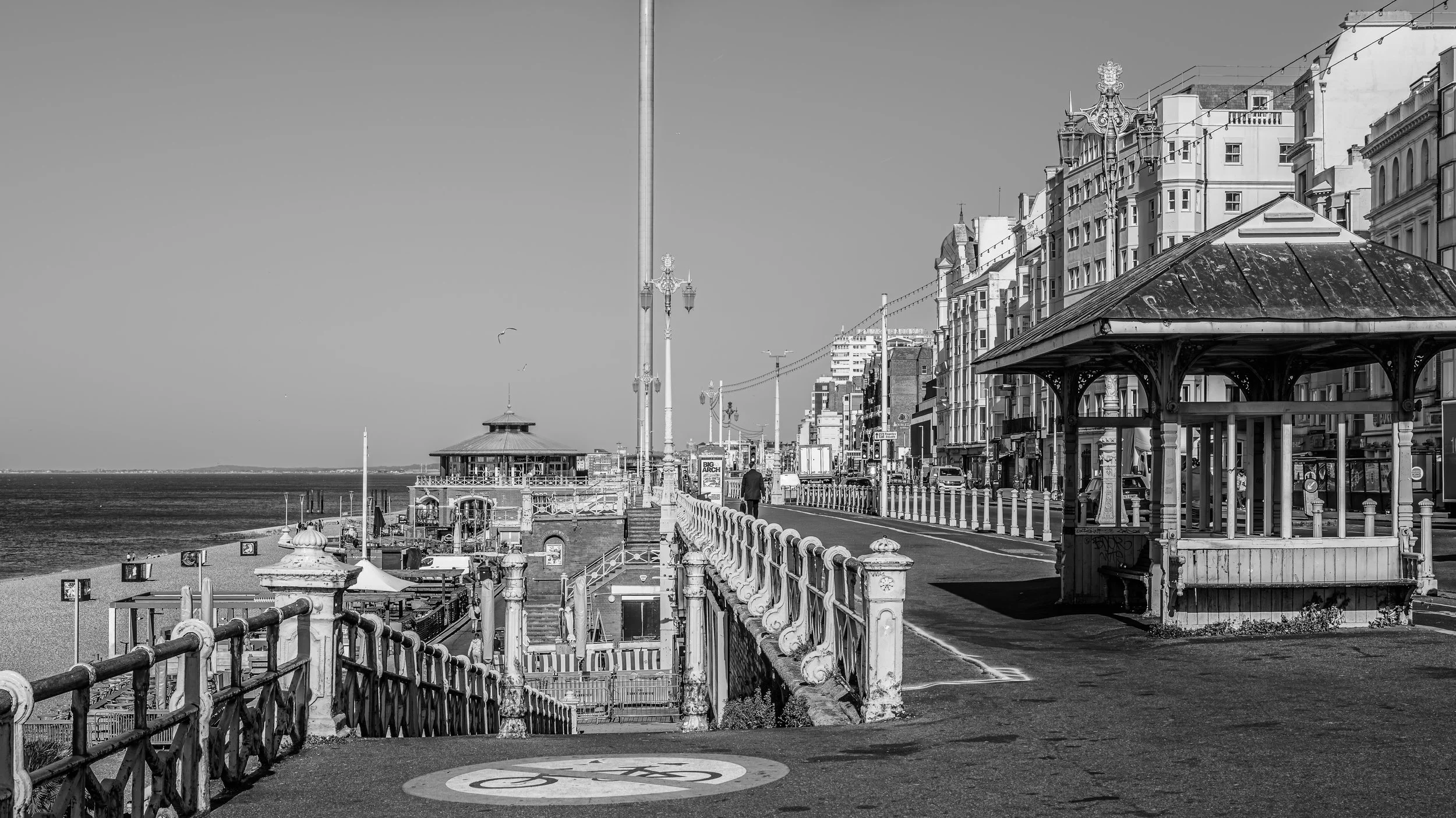 Black and white photo of a seaside boardwalk with Victorian-style architecture, street lamps, a gazebo-like structure, and a beach, showing a calm sea and a clear sky in the background.