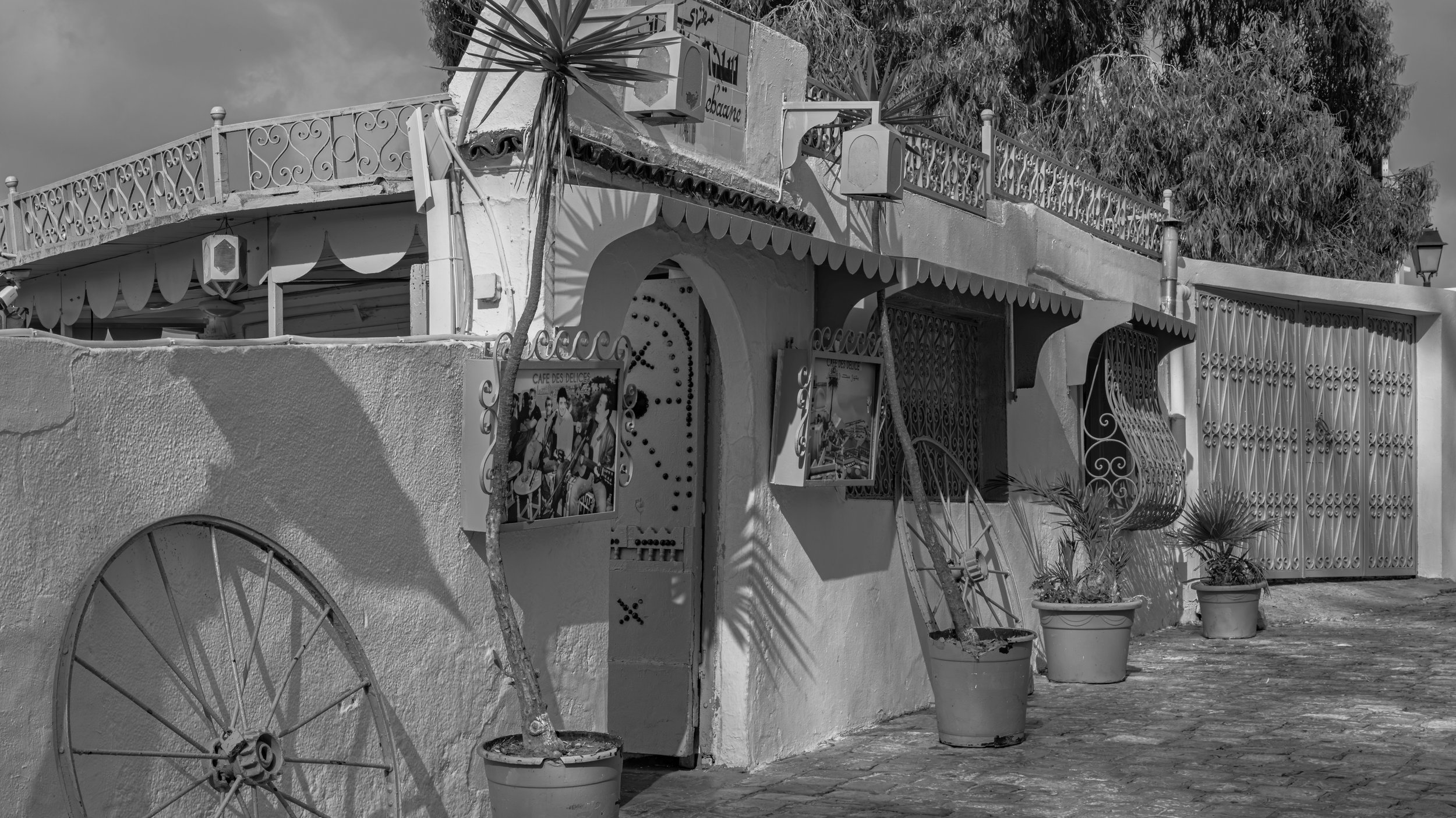 Black and white photo of a building with decorative iron balconies, potted plants outside, and a street lamp. A sign with the word 'ebacura' and a poster featuring musicians are visible on the building's exterior.