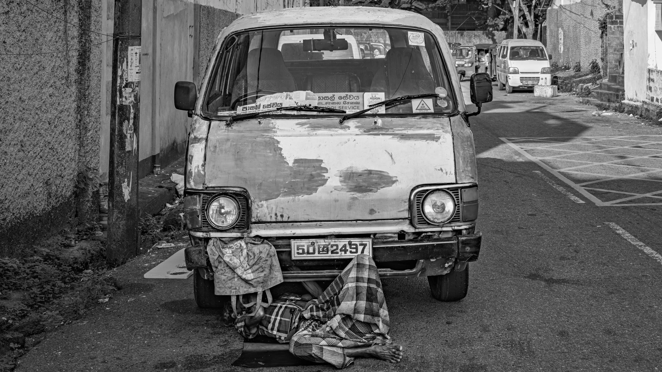An old, worn-out van parked on the side of a city street, with a person sleeping underneath it.
