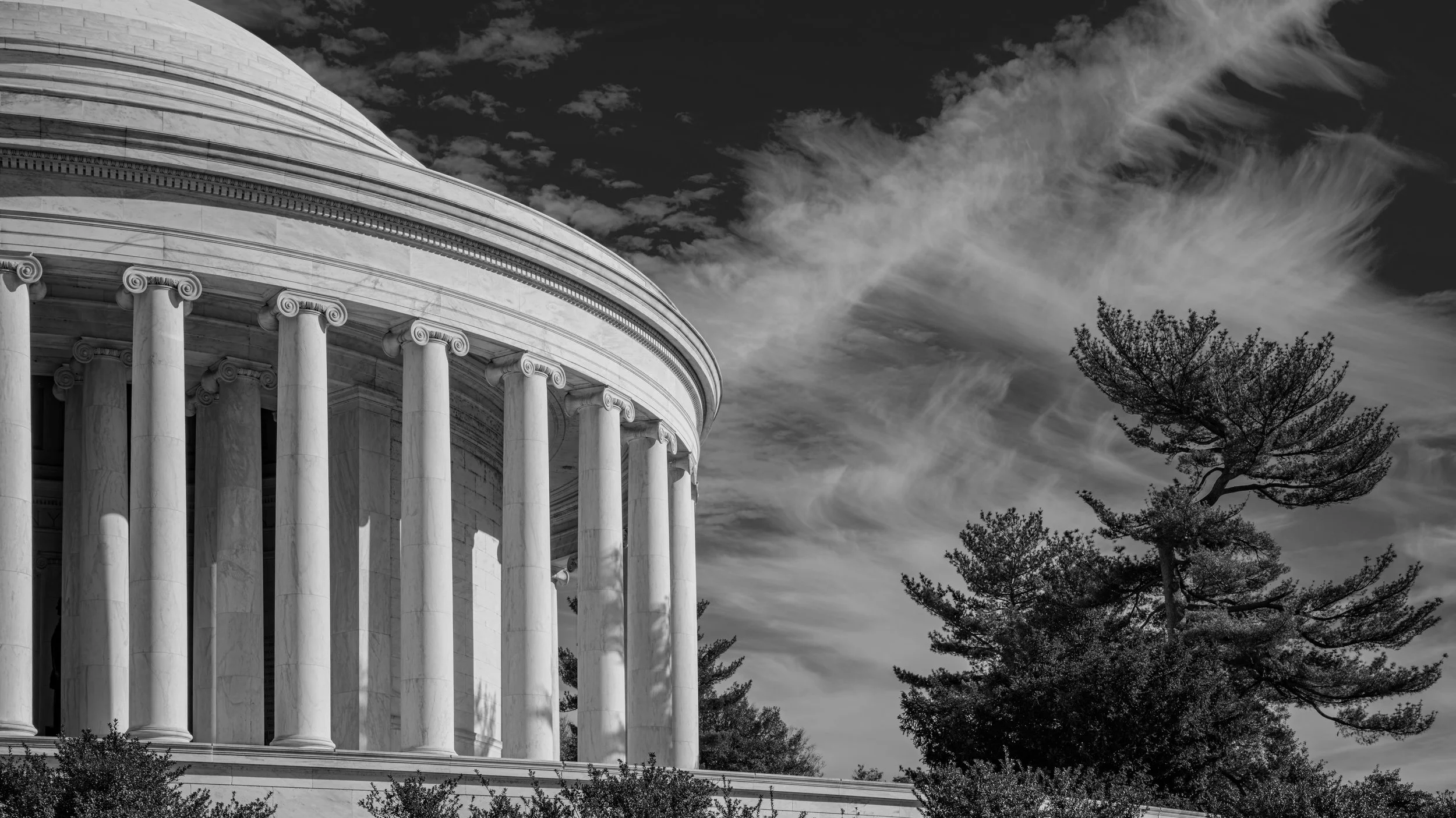 A black-and-white photo of the U.S. Supreme Court building with large columns and a domed roof, with clouds and trees in the background.