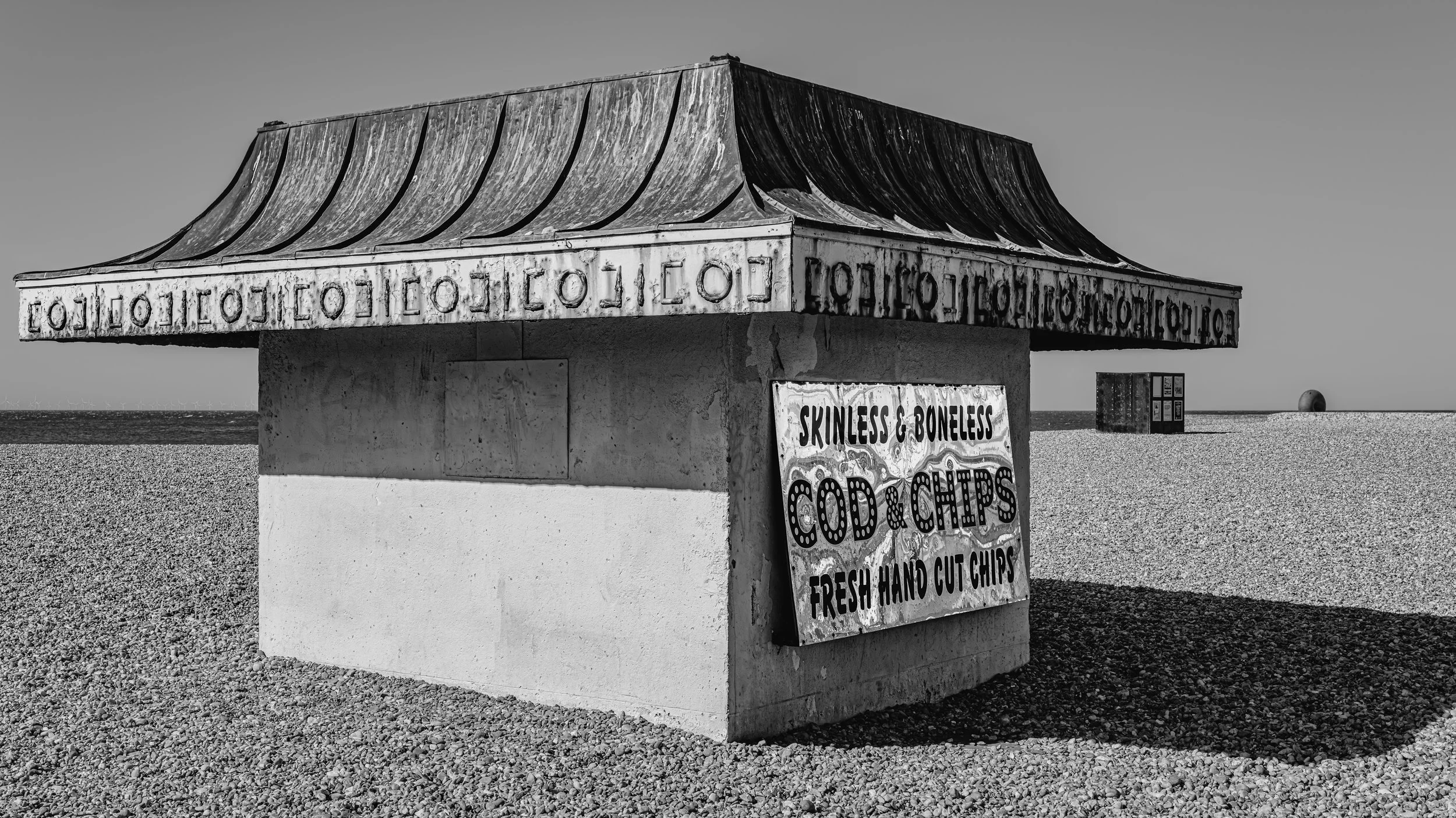 A small weathered wooden structure on a pebble-covered beach with a sign advertising seafood and chips, and a clear sky in the background.
