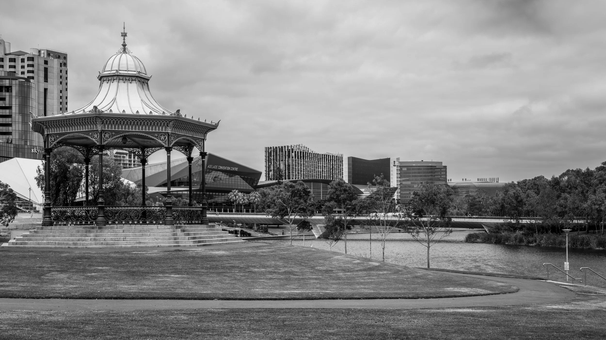 A cityscape featuring a historic ornate gazebo in the foreground, a body of water, modern high-rise buildings, and the Adelaide Convention Centre in the background, under a cloudy sky.