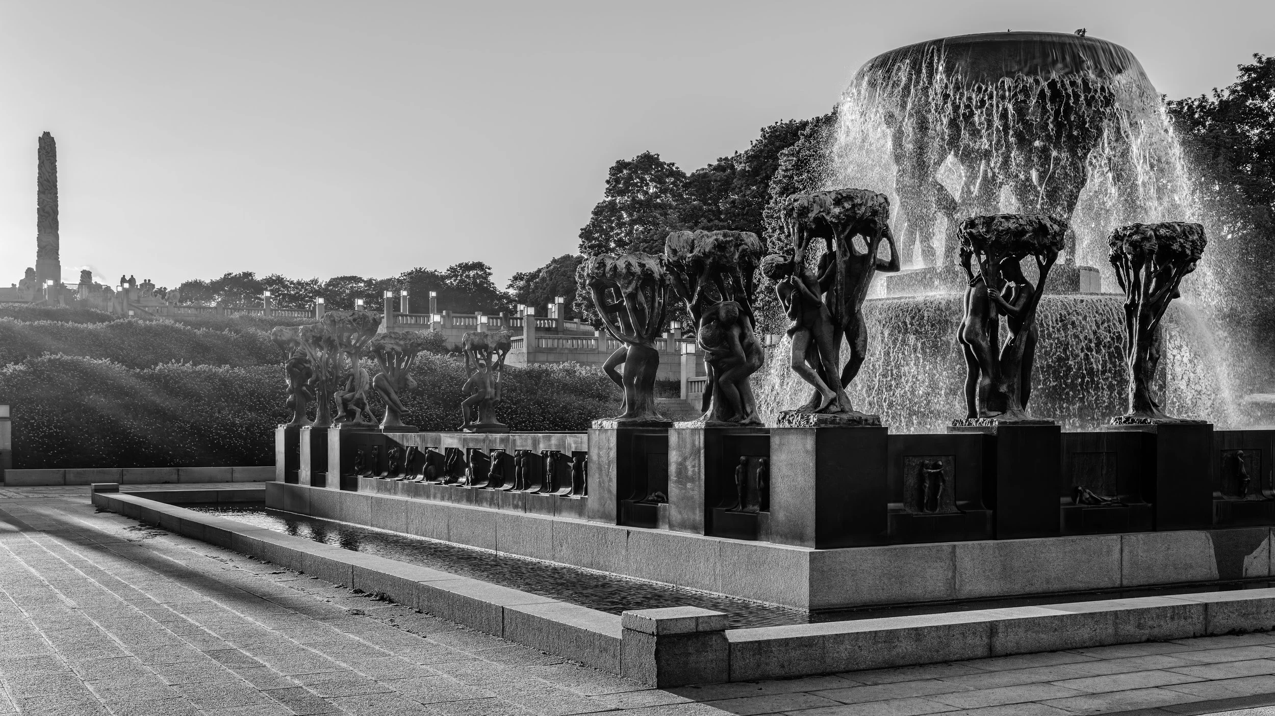 Black and white photo of a large fountain with statues of women holding large bowls from which water flows, with trees and a monument with a tall obelisk in the background.