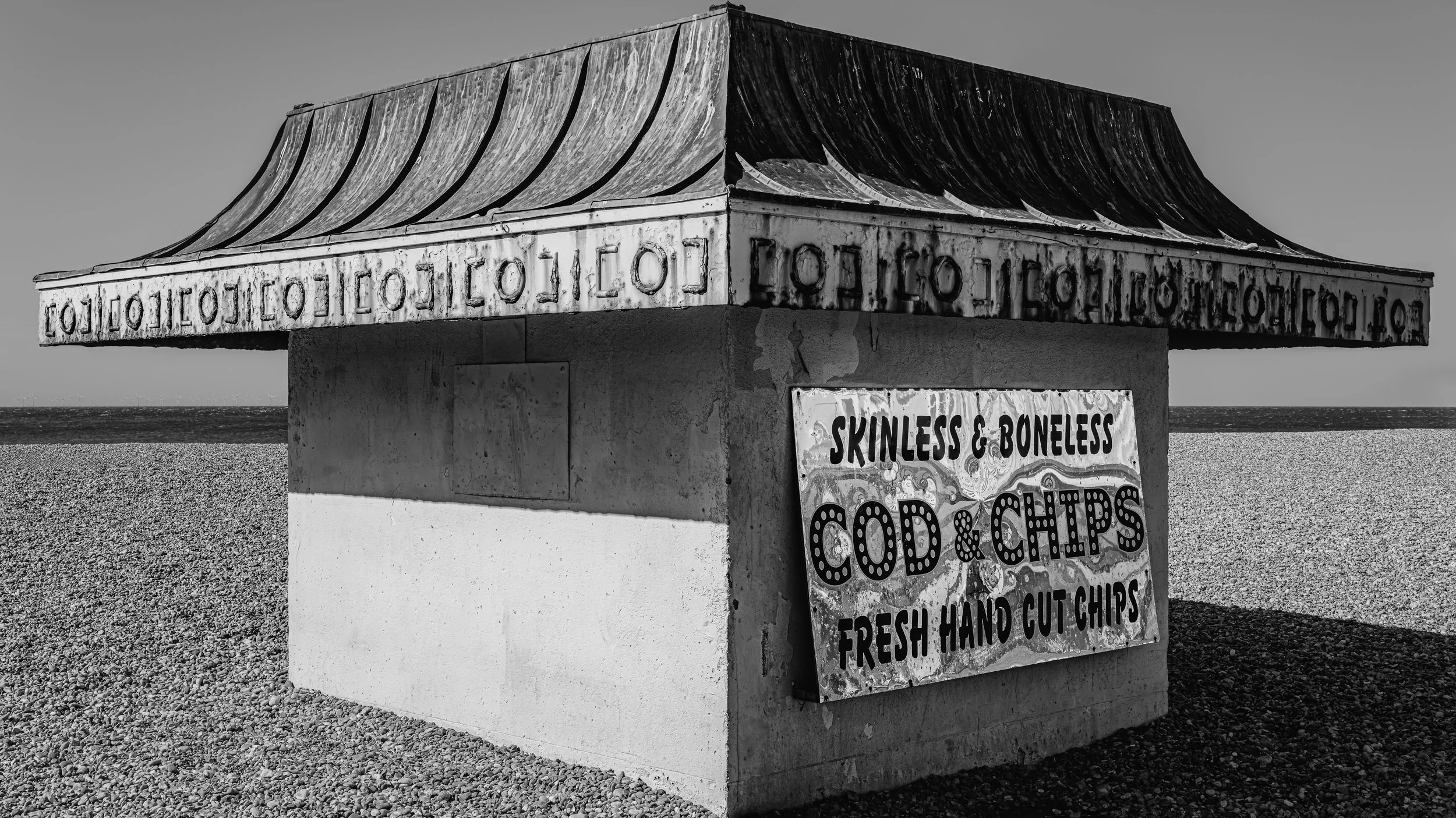 A small beachside food stand with a corrugated metal roof, advertising skinless and boneless cod and chips, and fresh hand-cut chips, located on a gravel beach with the ocean in the background. Brighton, England, UK.