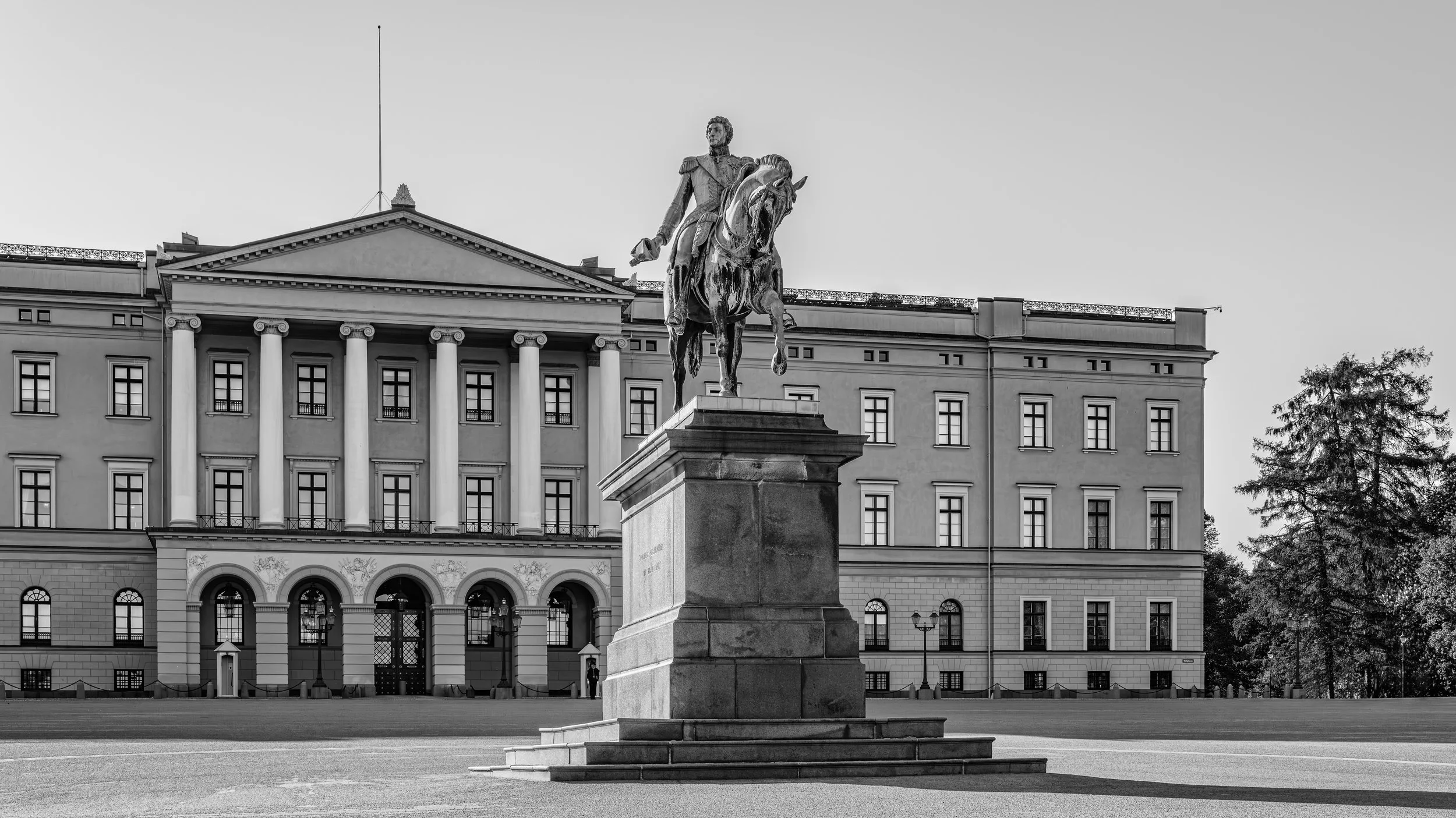 Black and white photo of a statue of a man on a horse in front of a grand building with columns and multiple windows, with trees to the right.