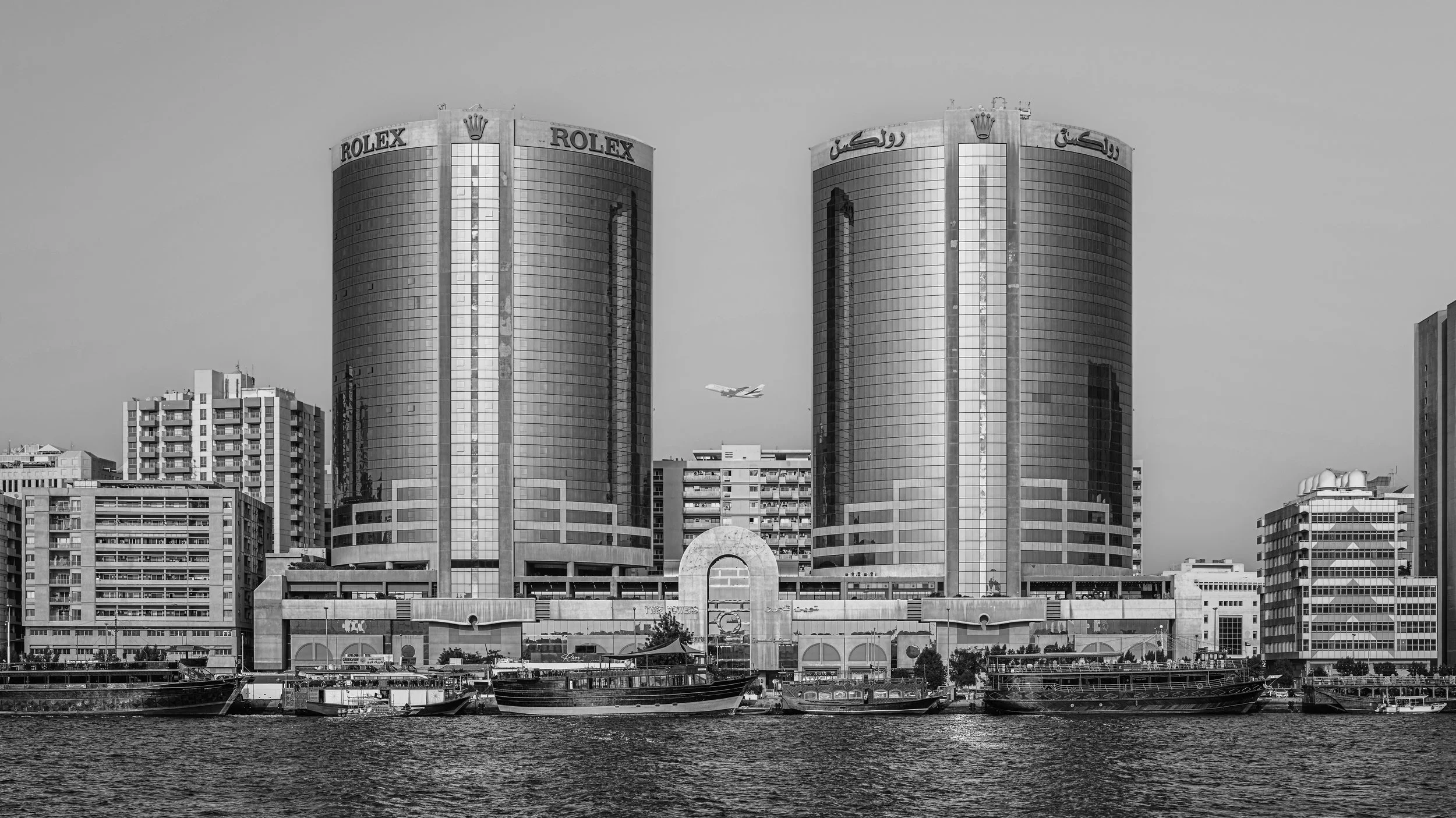Black and white cityscape featuring modern skyscrapers near water with boats, including a tall building with Rolex and Arabic signage.