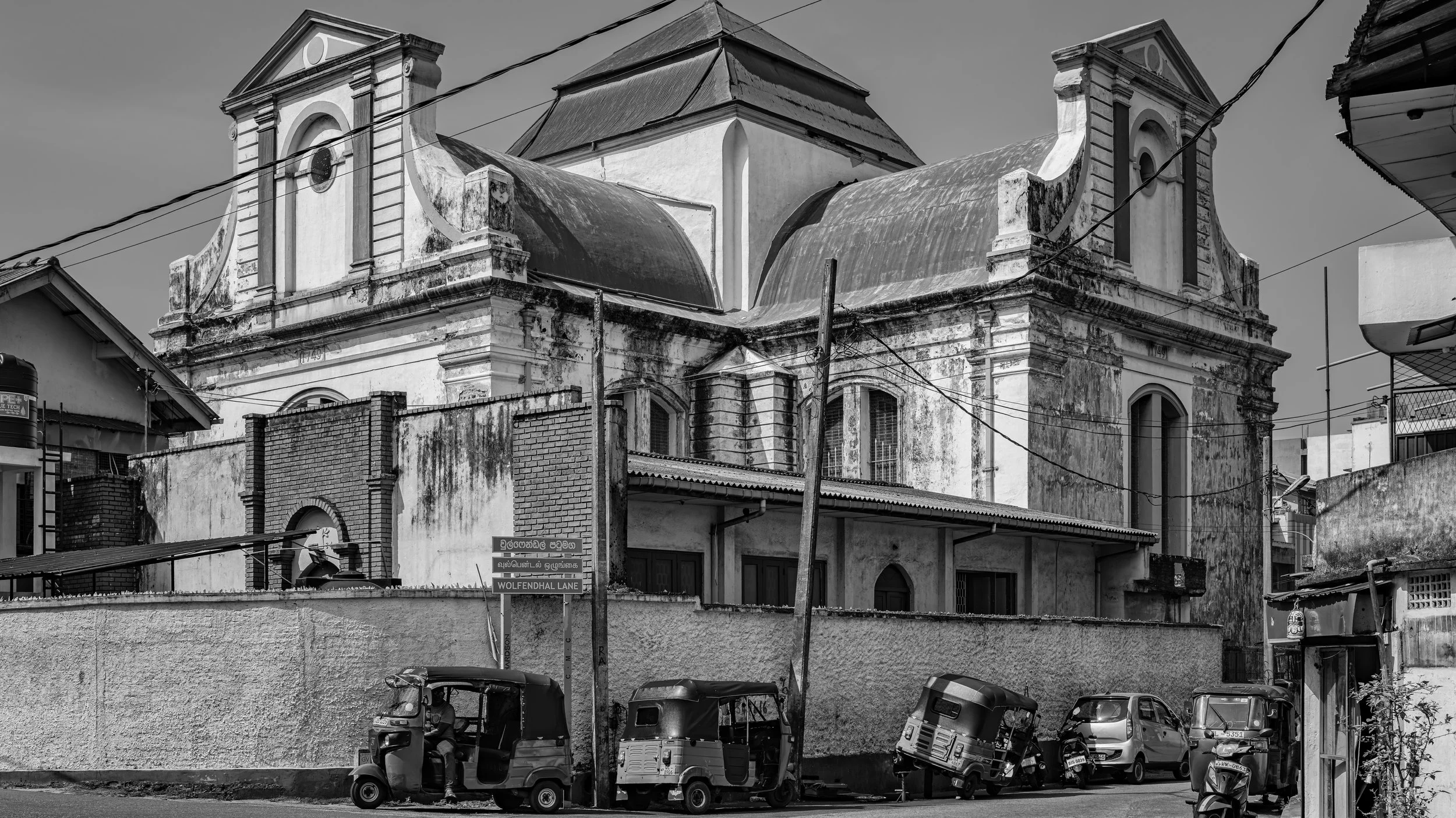 An old, worn church building with ornate architecture in black and white. Several vehicles, including three auto rickshaws and cars, are parked in front. Power lines cross over the scene, and street signs indicate the location as Wolfendahl Lane.