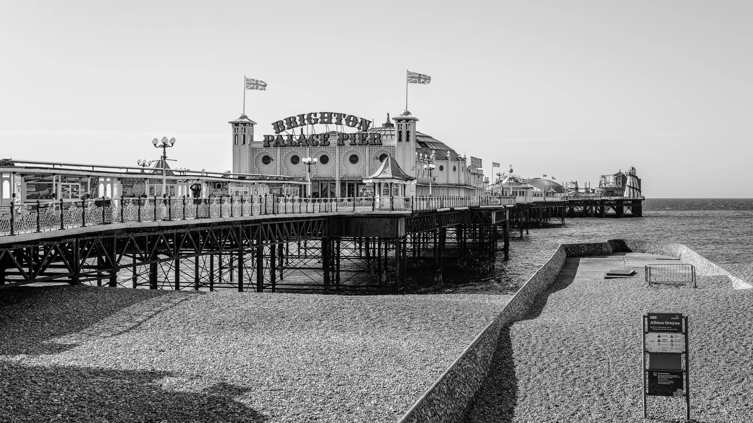 Black and white photo of Brighton Palace Pier extending over the sea with a cloudy sky, featuring a sign for Albion Grove at the foreground.