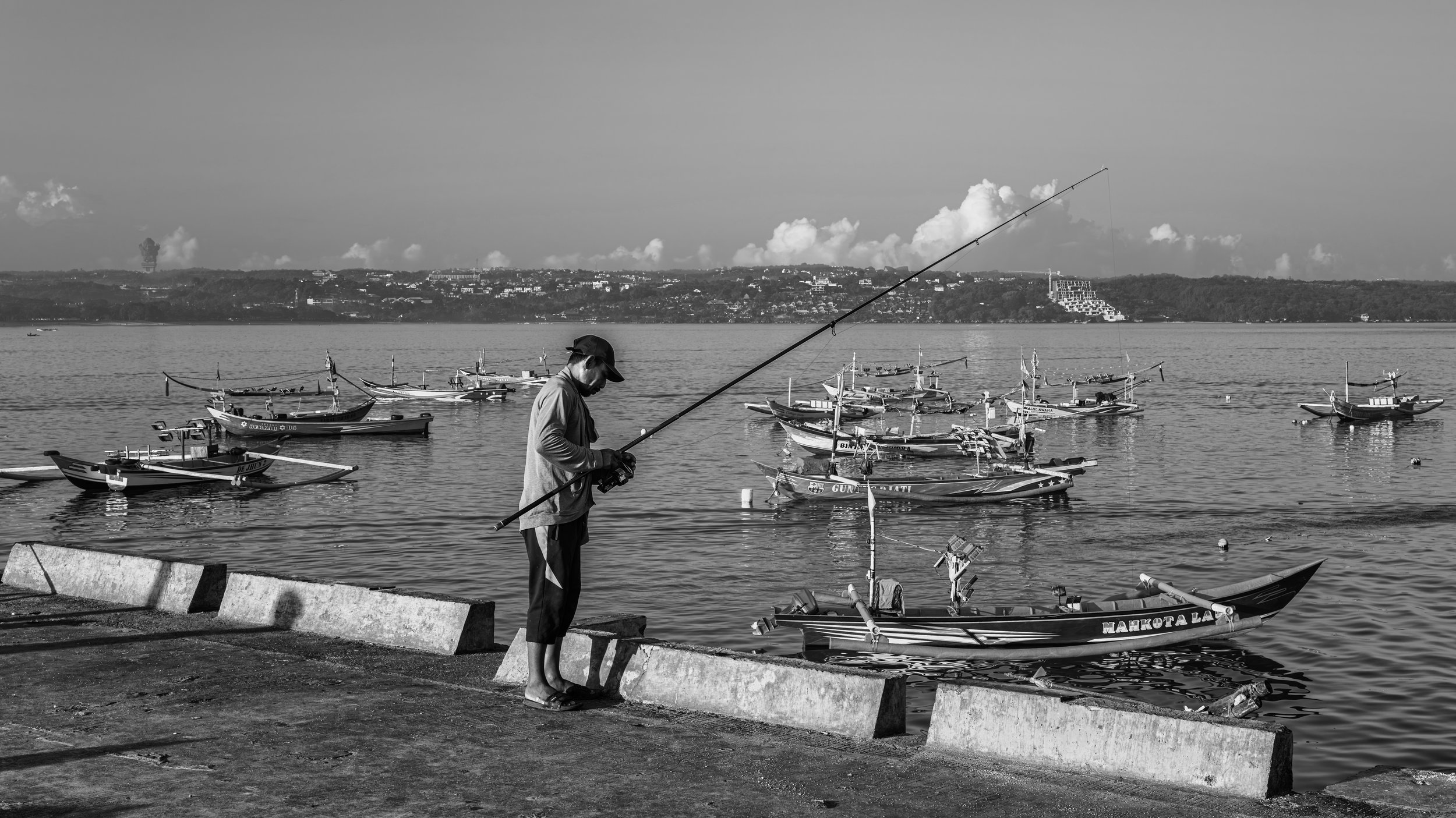 A man standing on a concrete ledge near the water, holding a fishing rod, with several small boats docked on the water behind him and a distant hilly landscape in the background.