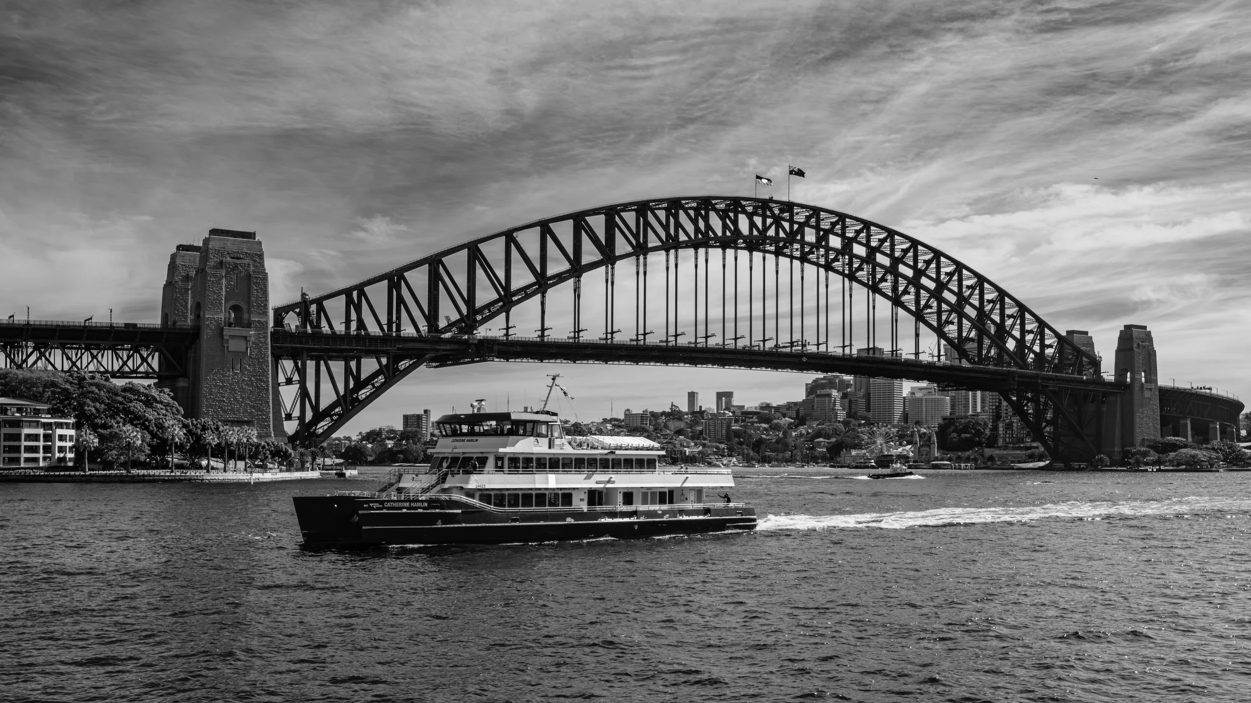 A black and white photo of Sydney Harbour Bridge with a boat named 'Catherine Hamlin' on the water below and city buildings in the background.
